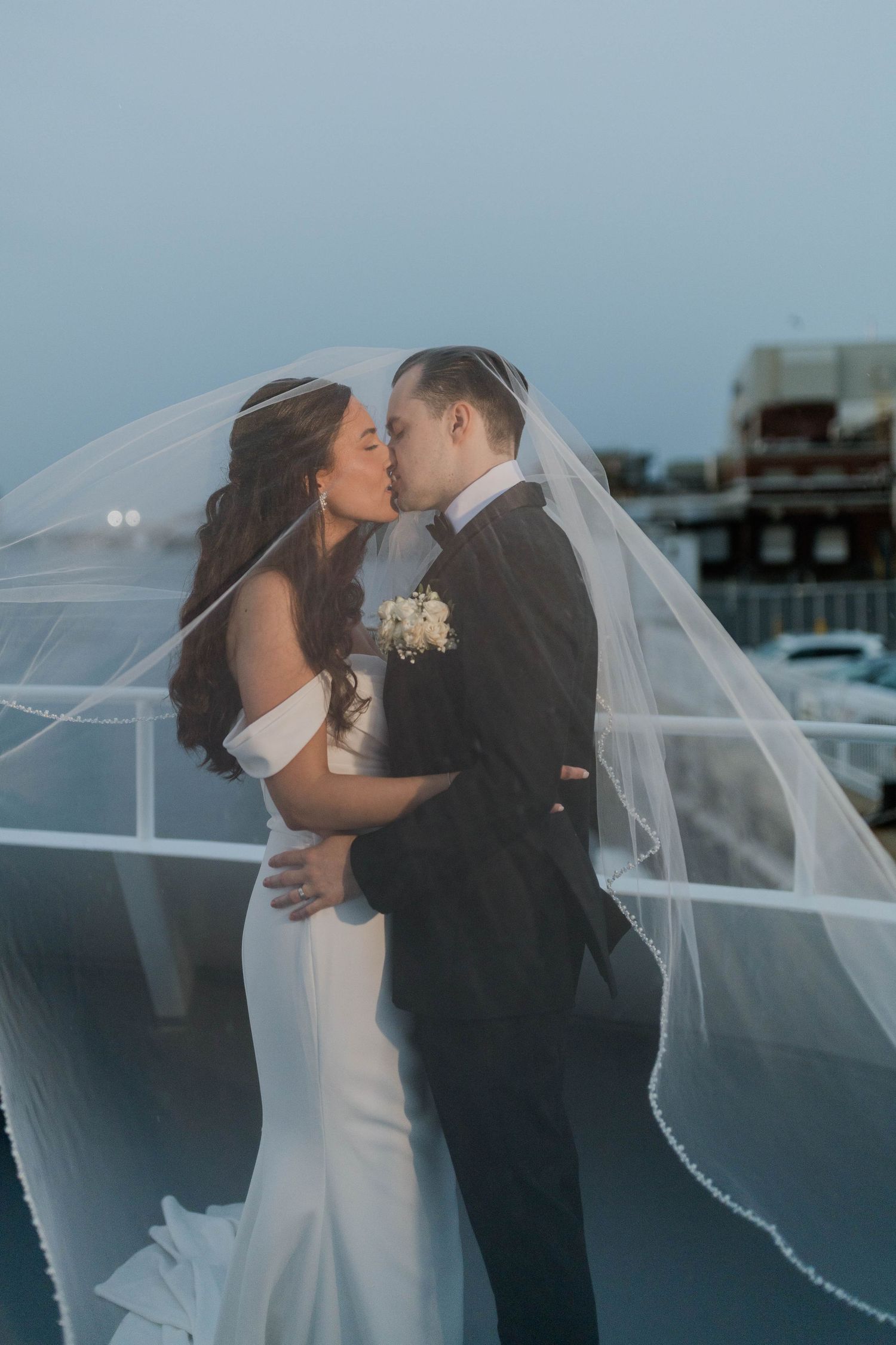 Bride and groom share romantic kiss outdoors at dusk with veil flowing in the wind.