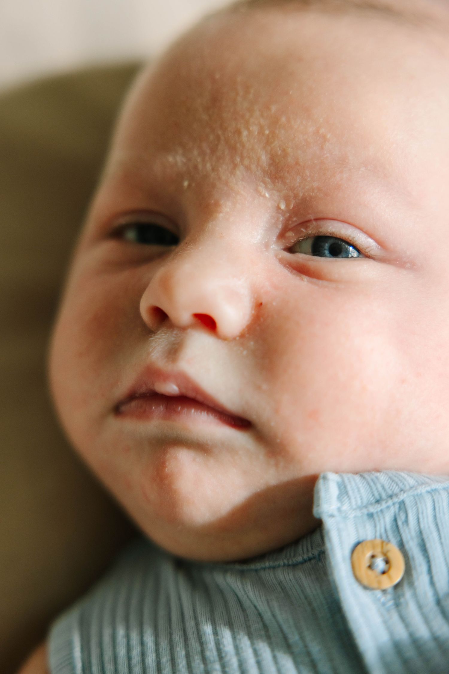 Close-up portrait of a newborn baby with blue eyes wearing a light blue ribbed onesie with button details.