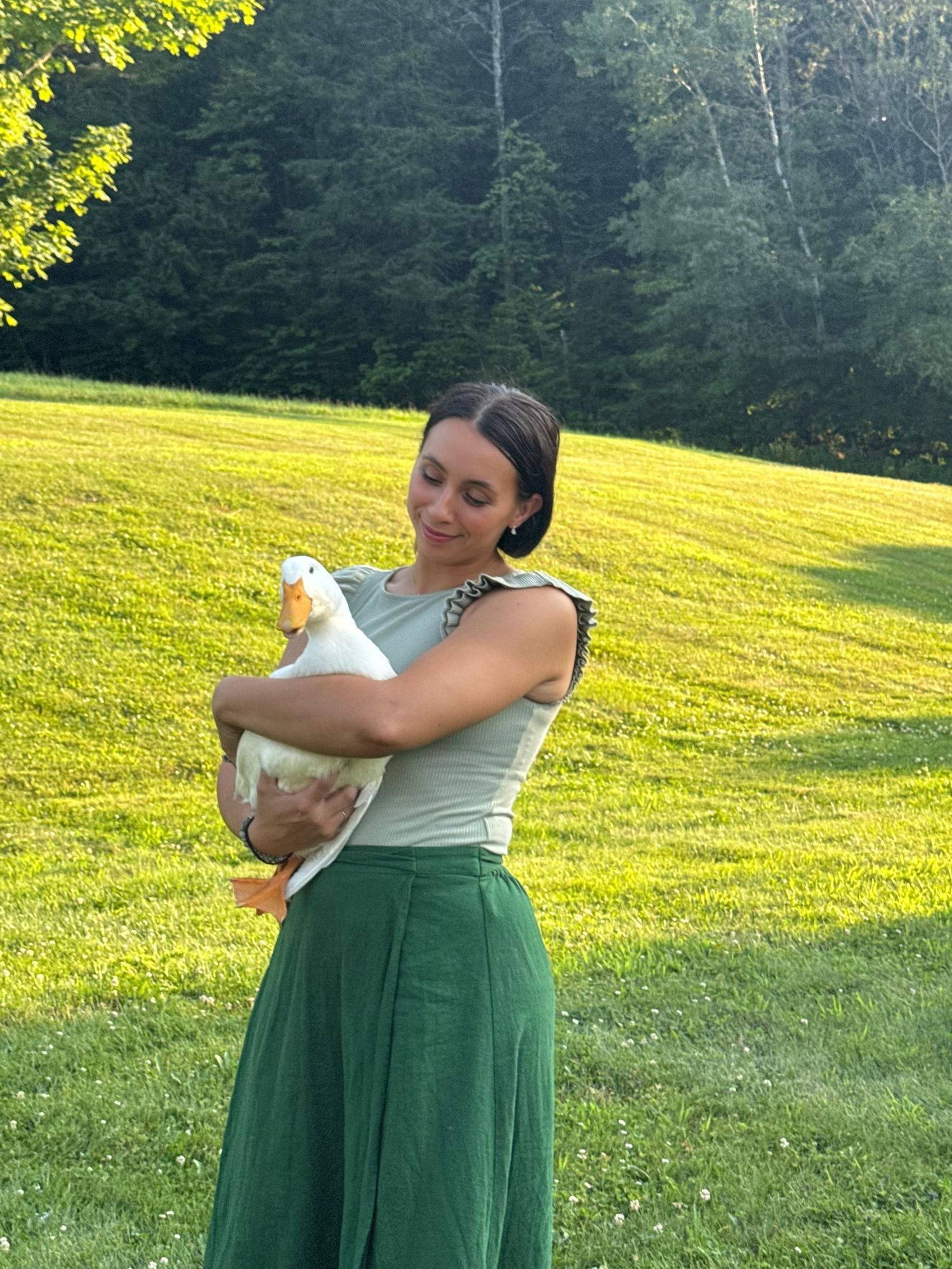 Woman in green skirt holding a white duck outdoors on a sunny green lawn with trees in background.