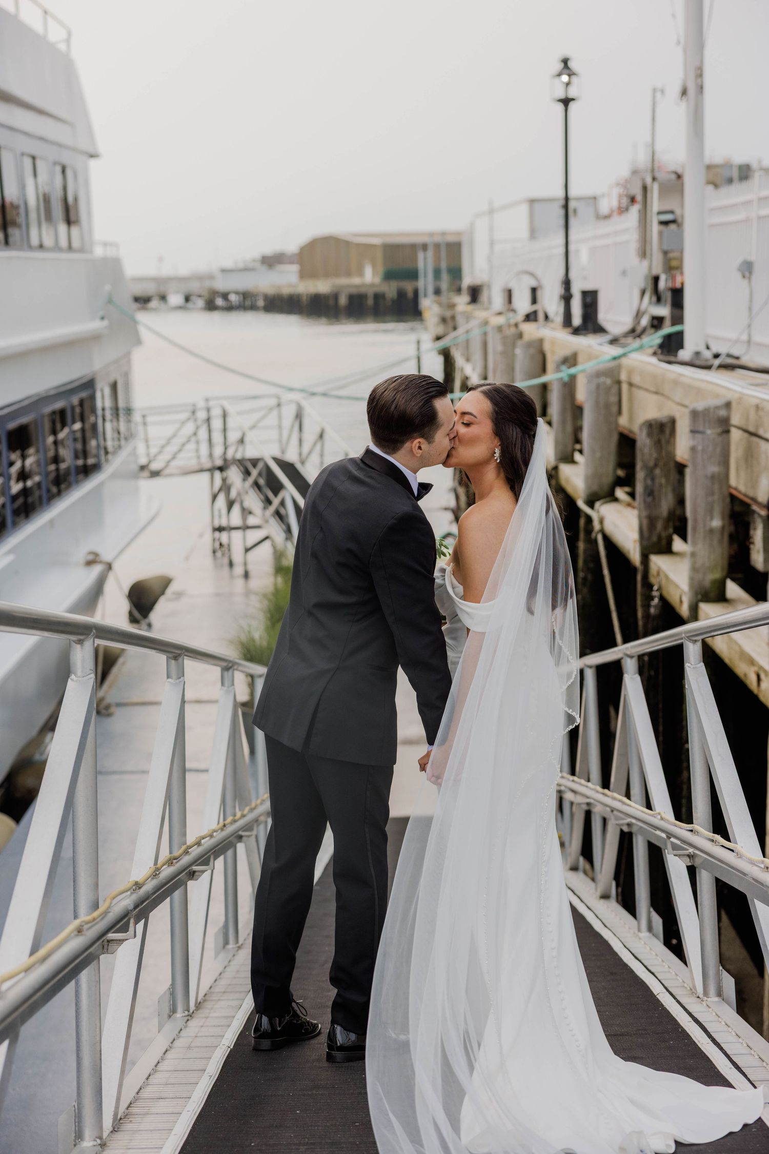 Bride and groom sharing a romantic kiss on a marina walkway, bride in flowing white gown with long veil.
