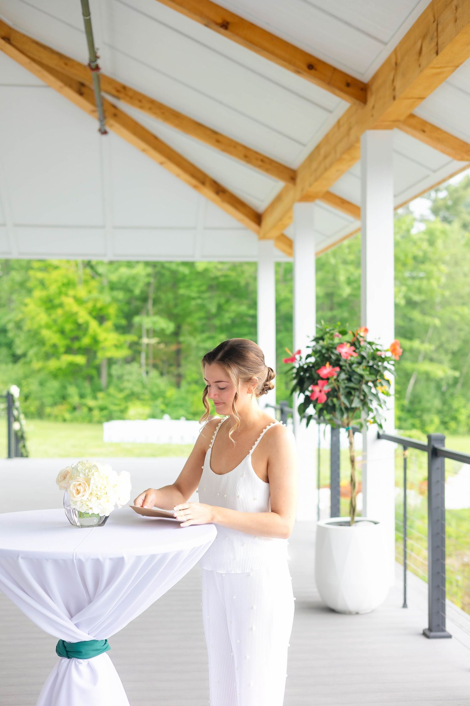 Bride in a white dress writing in a journal at a draped cocktail table on a bright outdoor pavilion.