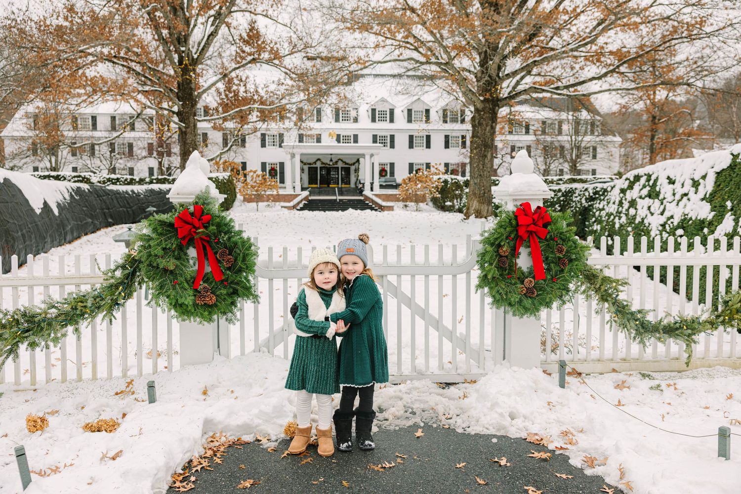 Two young sisters in green dresses hug near a festive white picket gate decorated with holiday wreaths in a snowy setting.