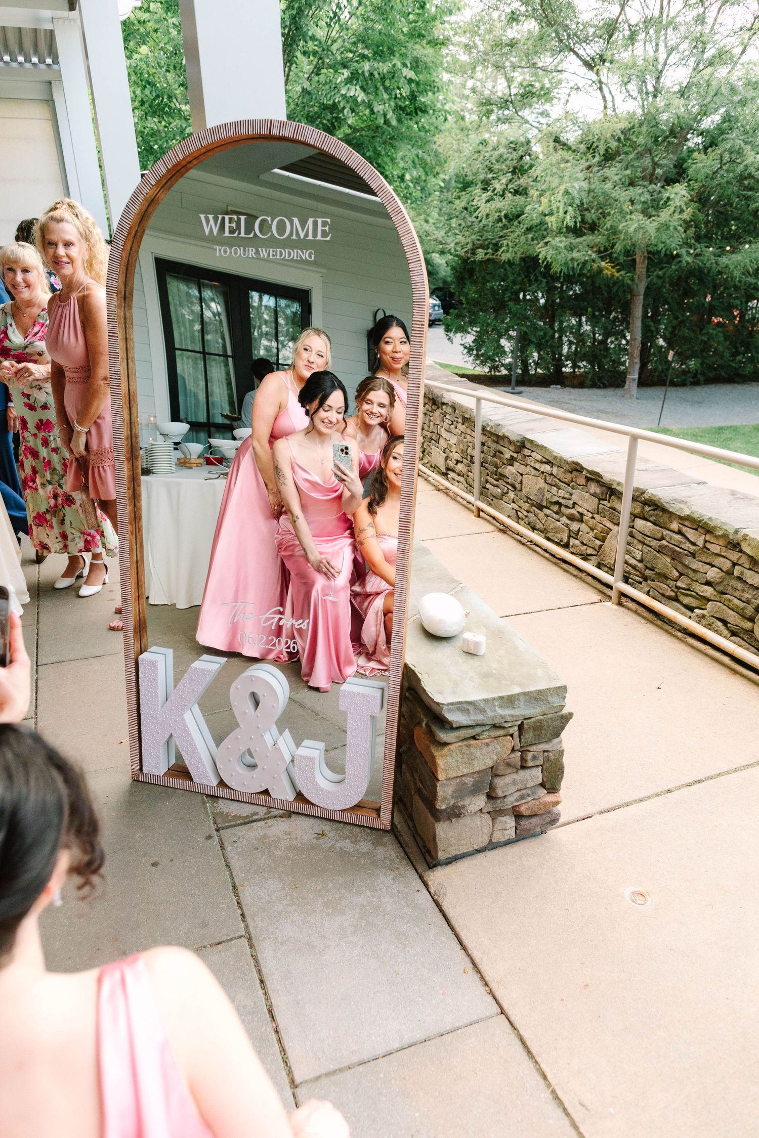Bridesmaids in pink dresses take a mirror selfie at K&J wedding welcome sign display.