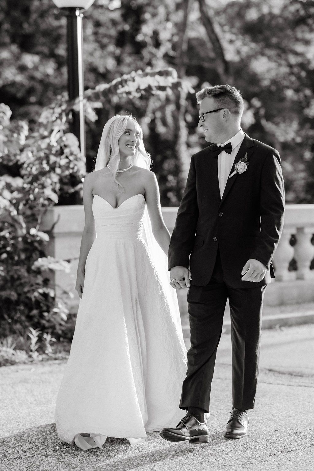 Black and white portrait of bride and groom holding hands and smiling while walking outdoors.