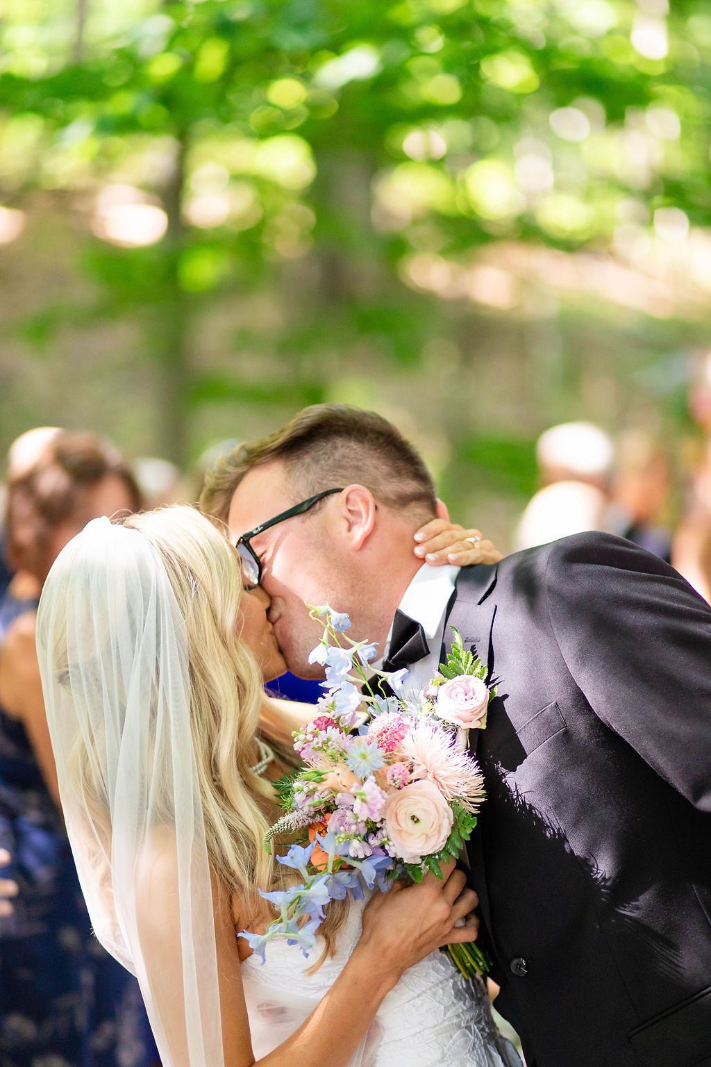 Bride and groom share first kiss at outdoor wedding ceremony surrounded by guests among green trees.