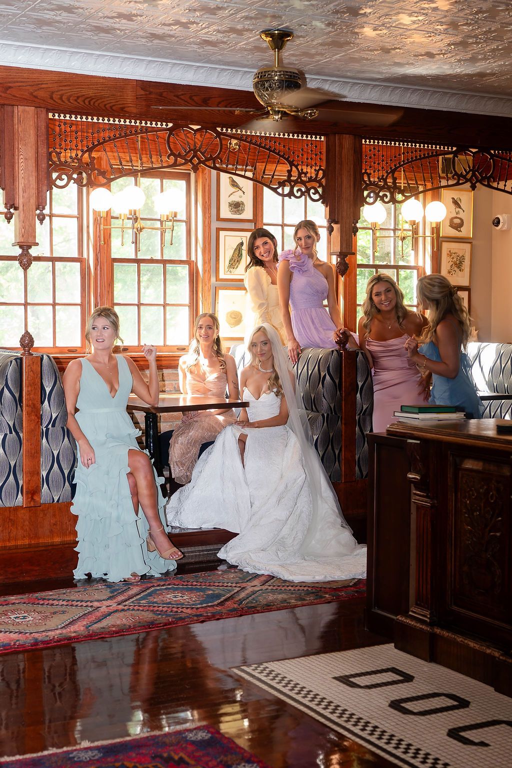 Bride in white gown poses with bridesmaids in pastel dresses inside a vintage wood-paneled room.