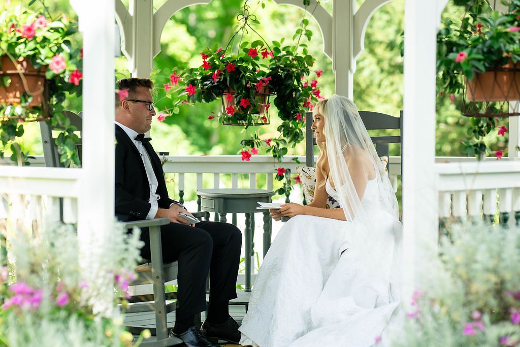Bride and groom share a quiet moment together in a flower-adorned white gazebo surrounded by colorful garden blooms.