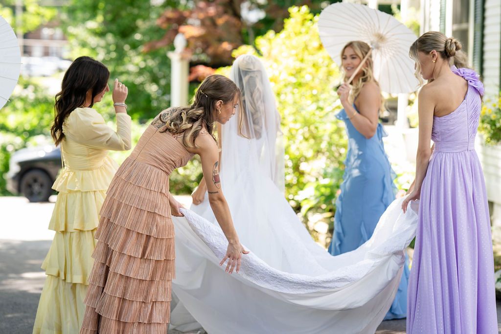 Bridesmaids in pastel dresses helping bride with her white wedding gown and veil outdoors.