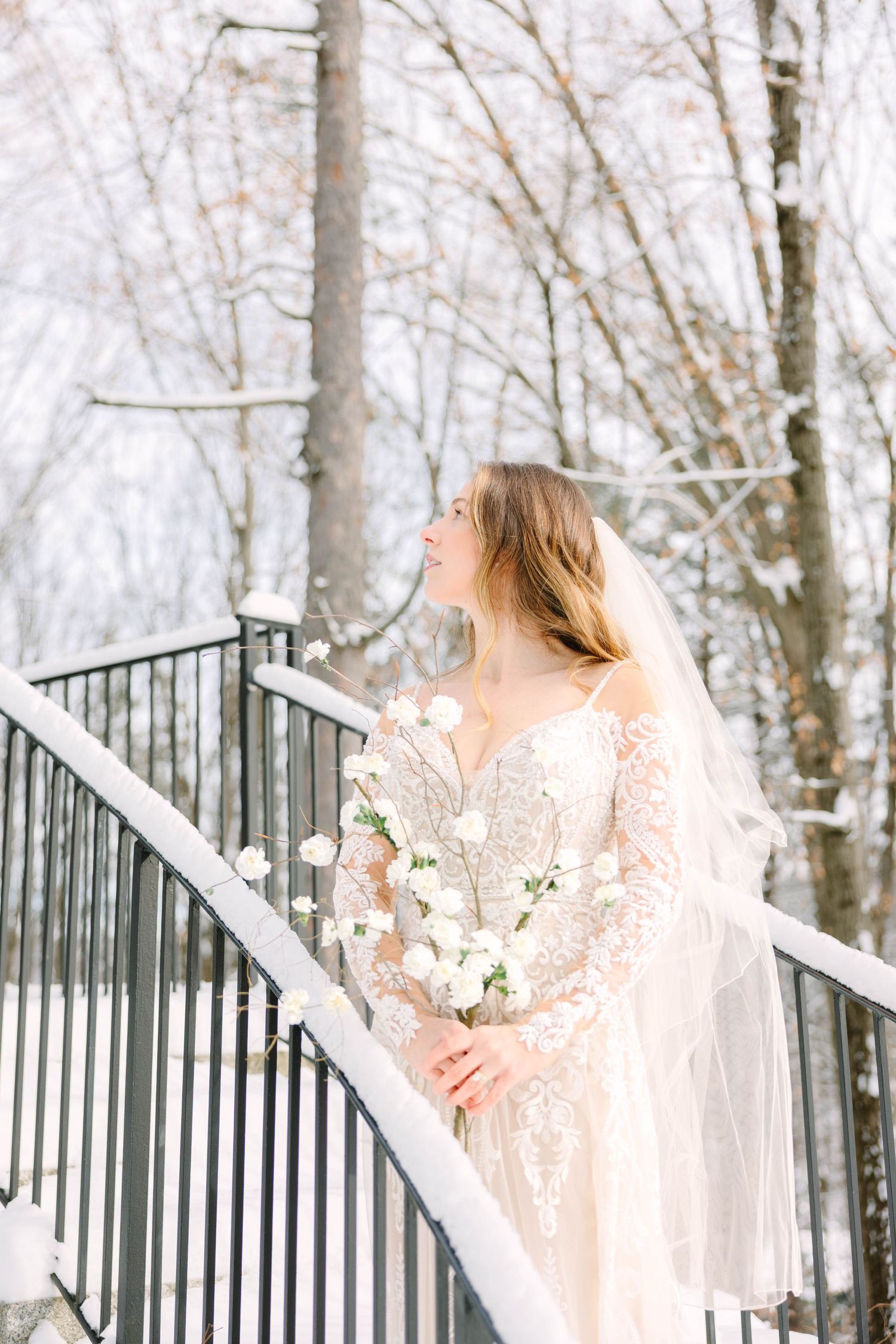 Bride in lace wedding dress and veil gazes upward on snowy staircase holding white flower bouquet.