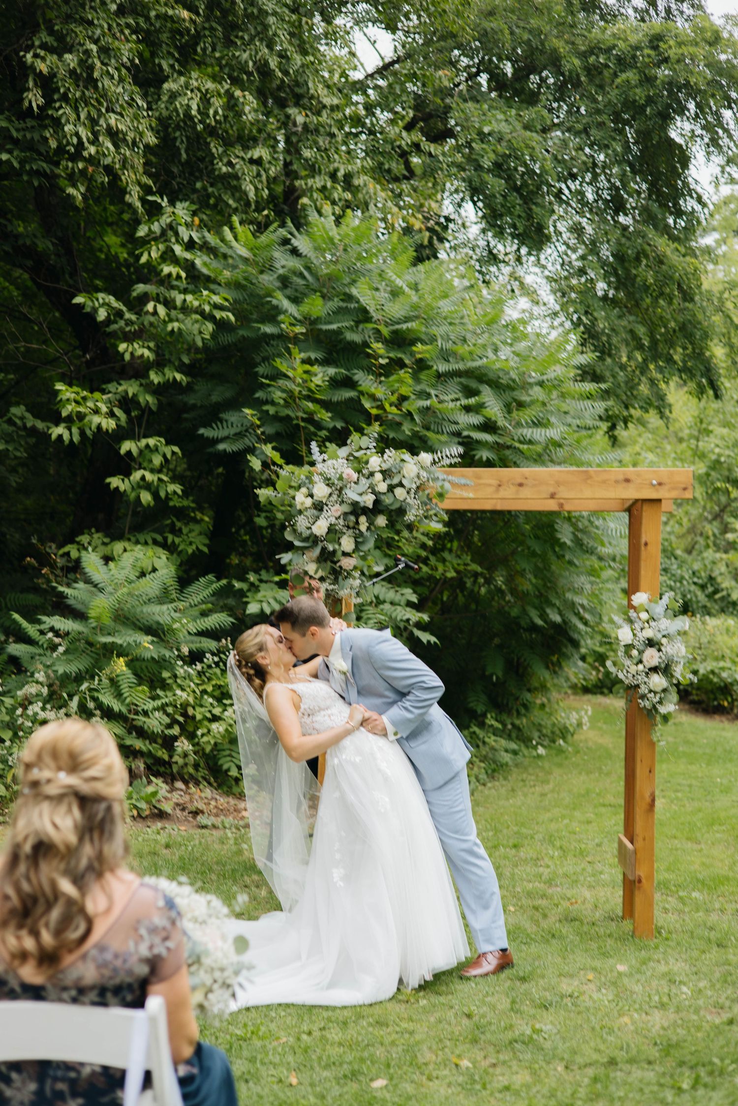 Bride and groom share first kiss under wooden arch with floral arrangements at outdoor garden wedding ceremony.