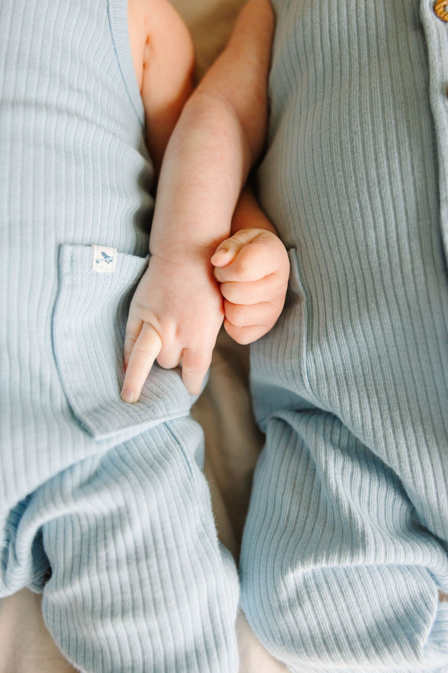 Two newborn twins in matching blue ribbed rompers holding hands together.