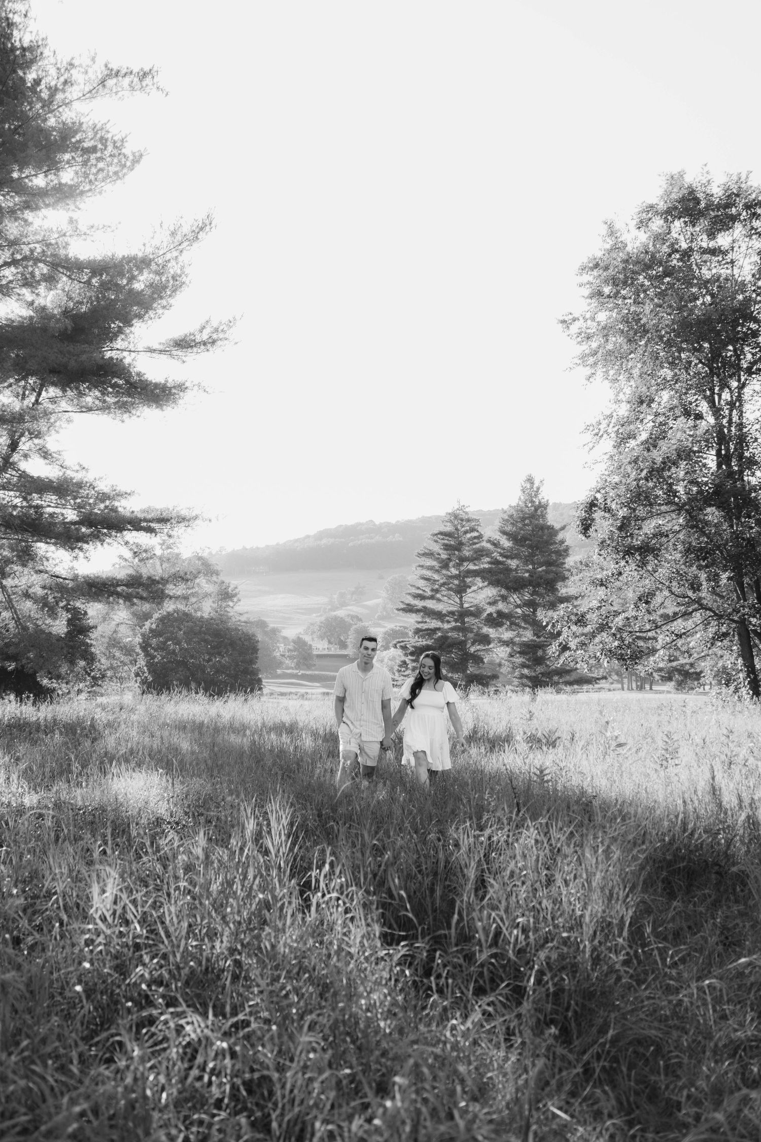 Black and white photo of a couple holding hands in a lush meadow surrounded by trees and rolling hills.