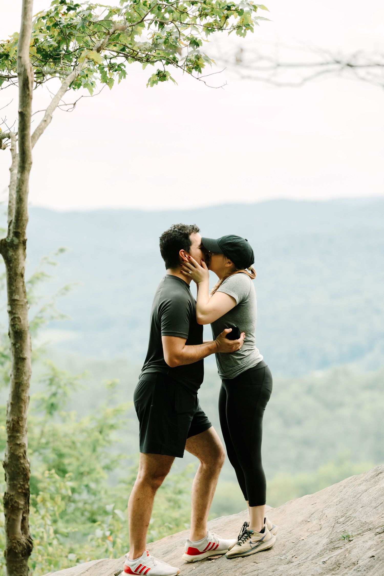 Couple sharing a romantic kiss on a rocky overlook surrounded by lush green forest and mountain views.