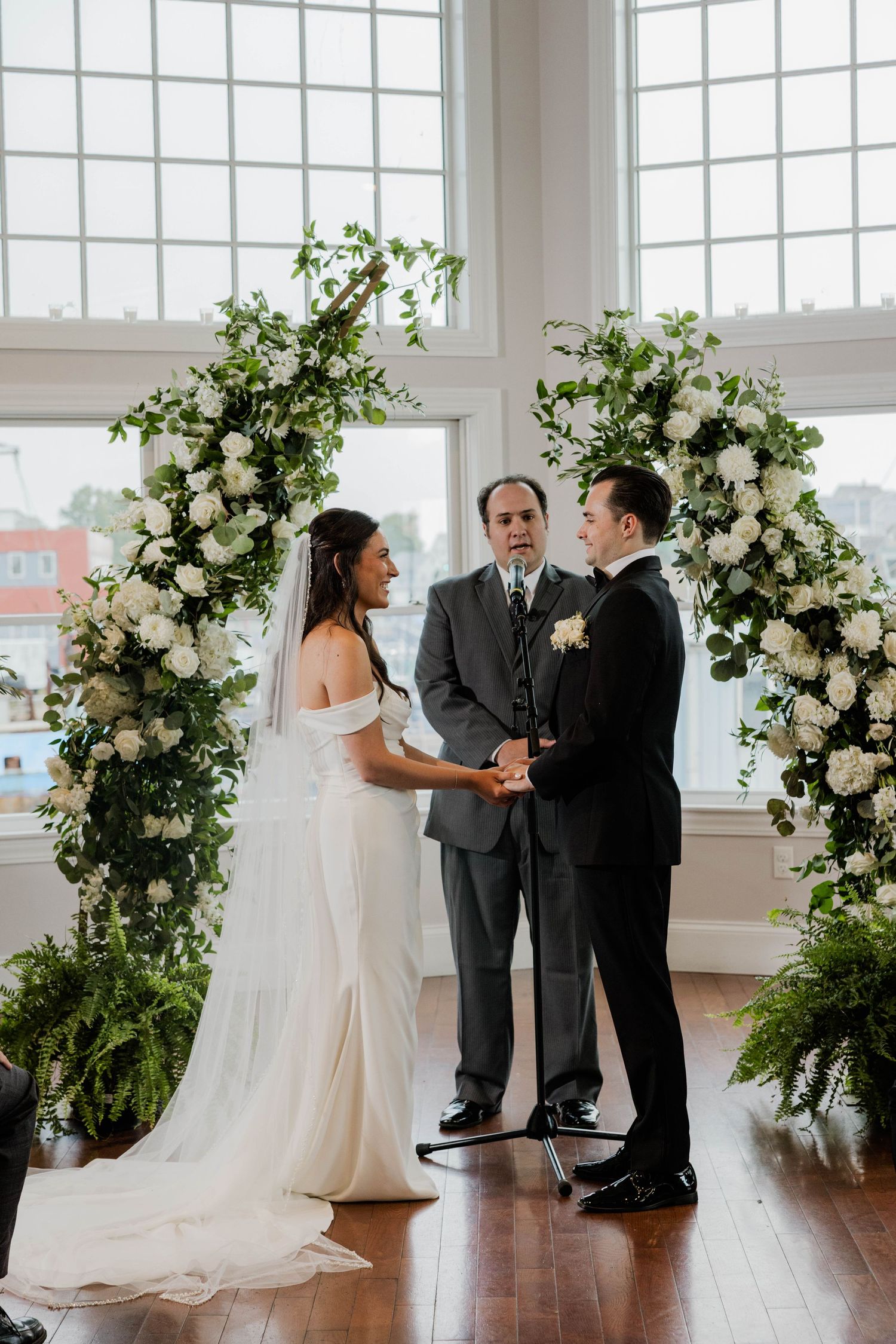 Bride and groom exchange vows under floral arch with white roses and greenery at indoor wedding.