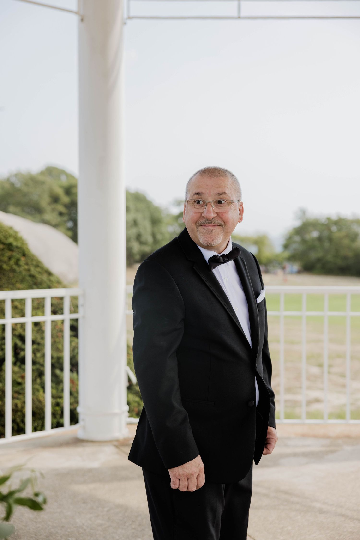 Groom in black tuxedo with bow tie smiling at a wedding venue gazebo.