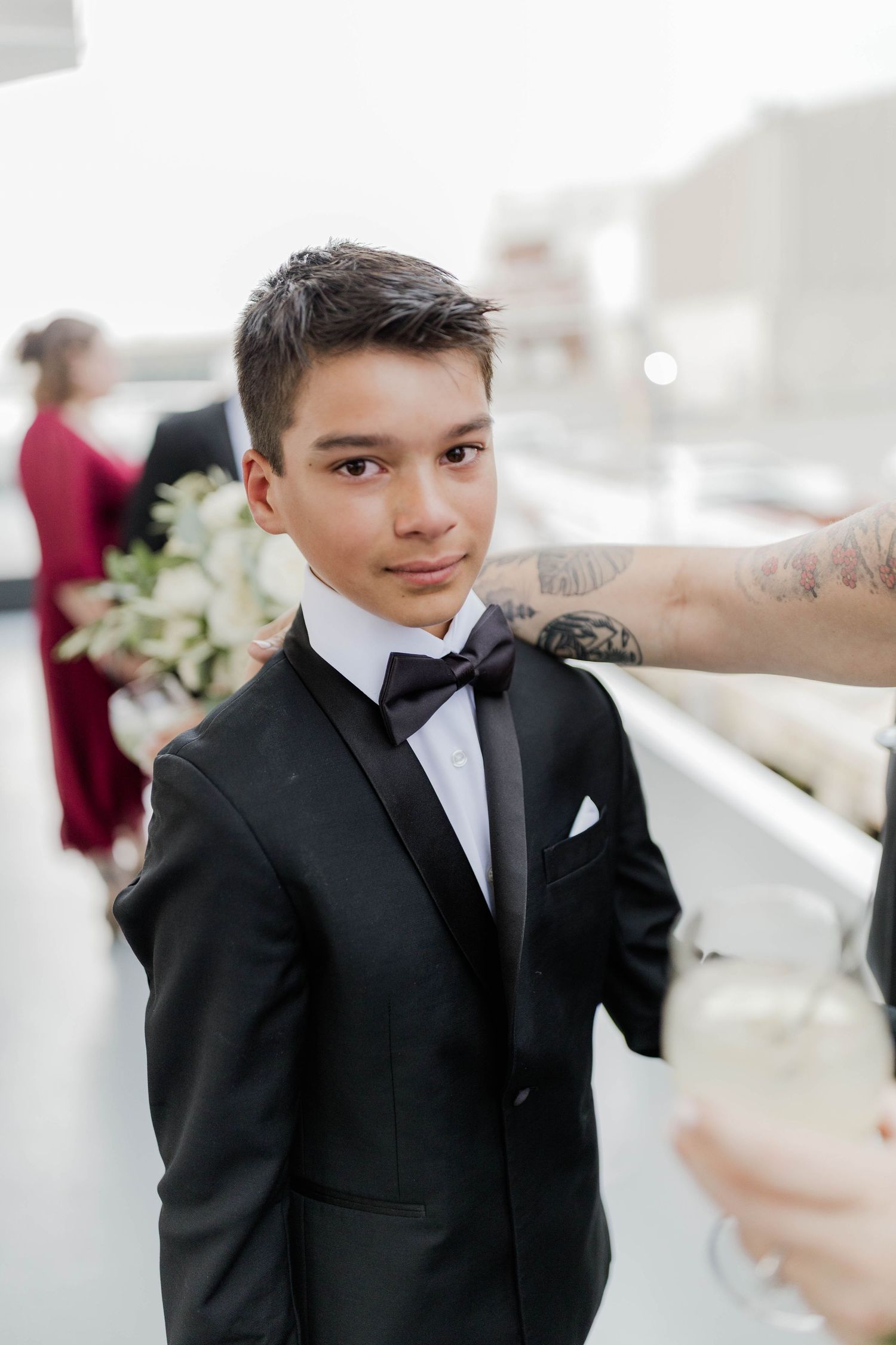 Young boy in a black tuxedo and bow tie posing at an outdoor wedding reception with champagne.