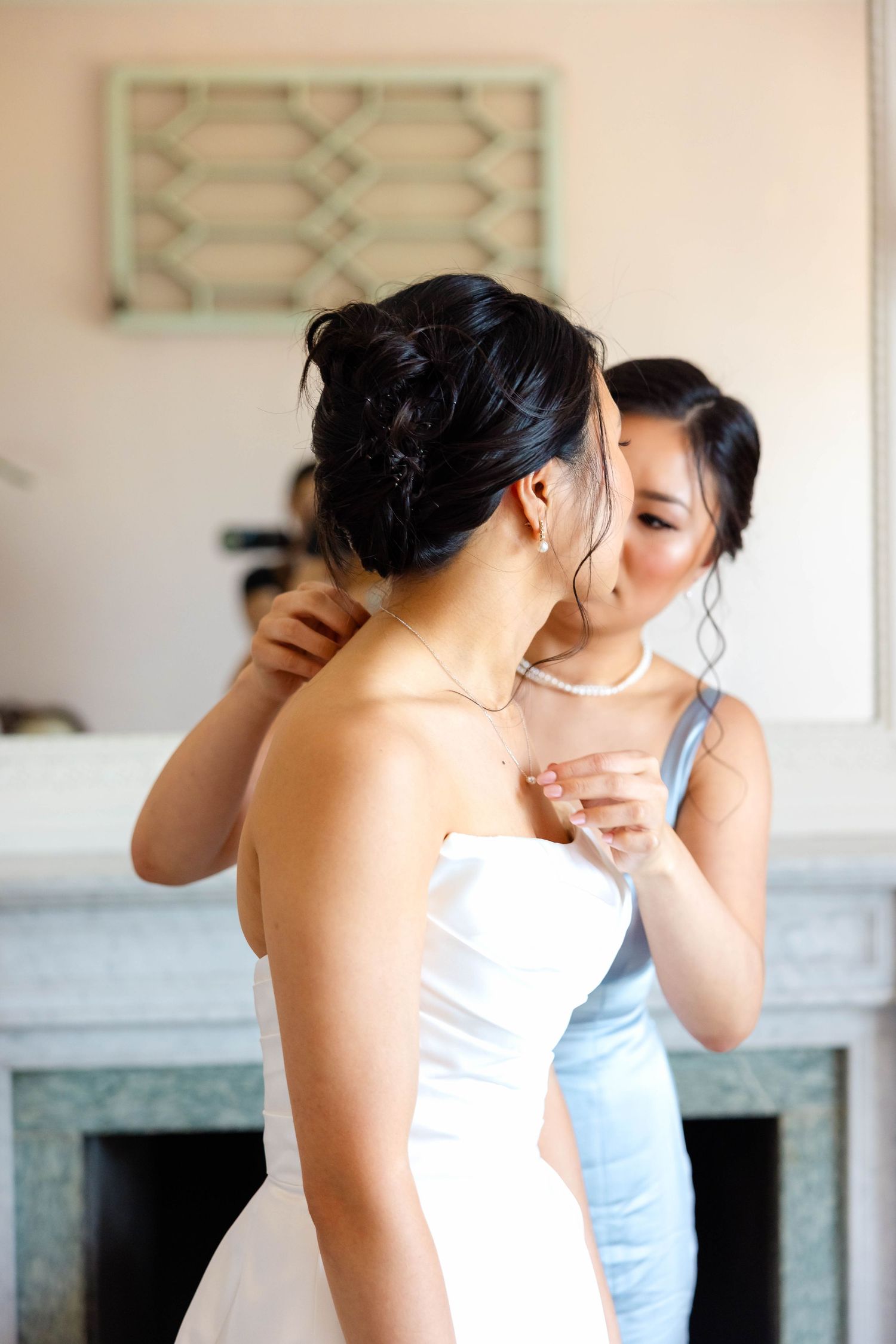 Bridesmaid in blue dress helping bride in white strapless gown with necklace before wedding ceremony.