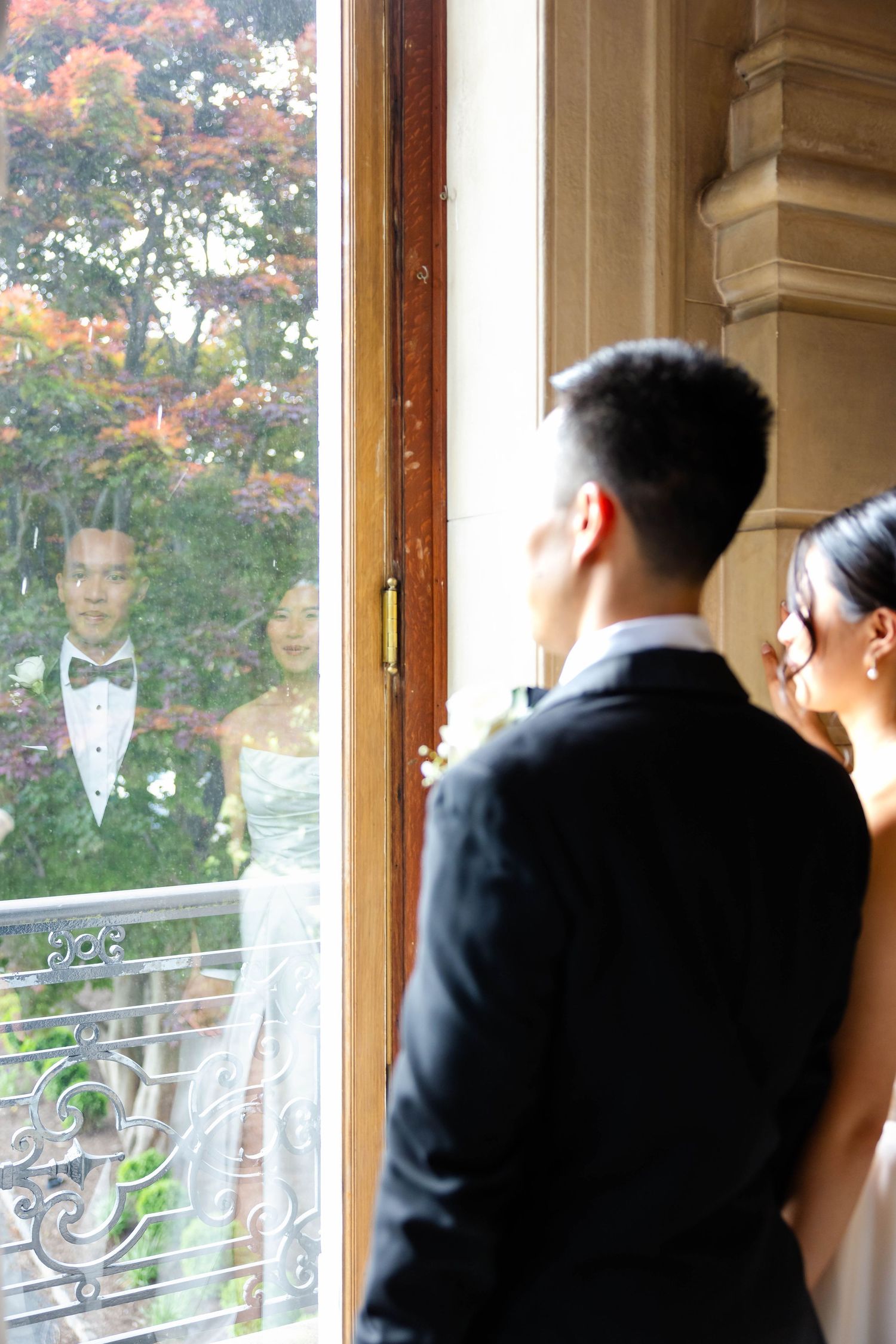Bride and groom reflected in ornate window with colorful autumn foliage visible outside.