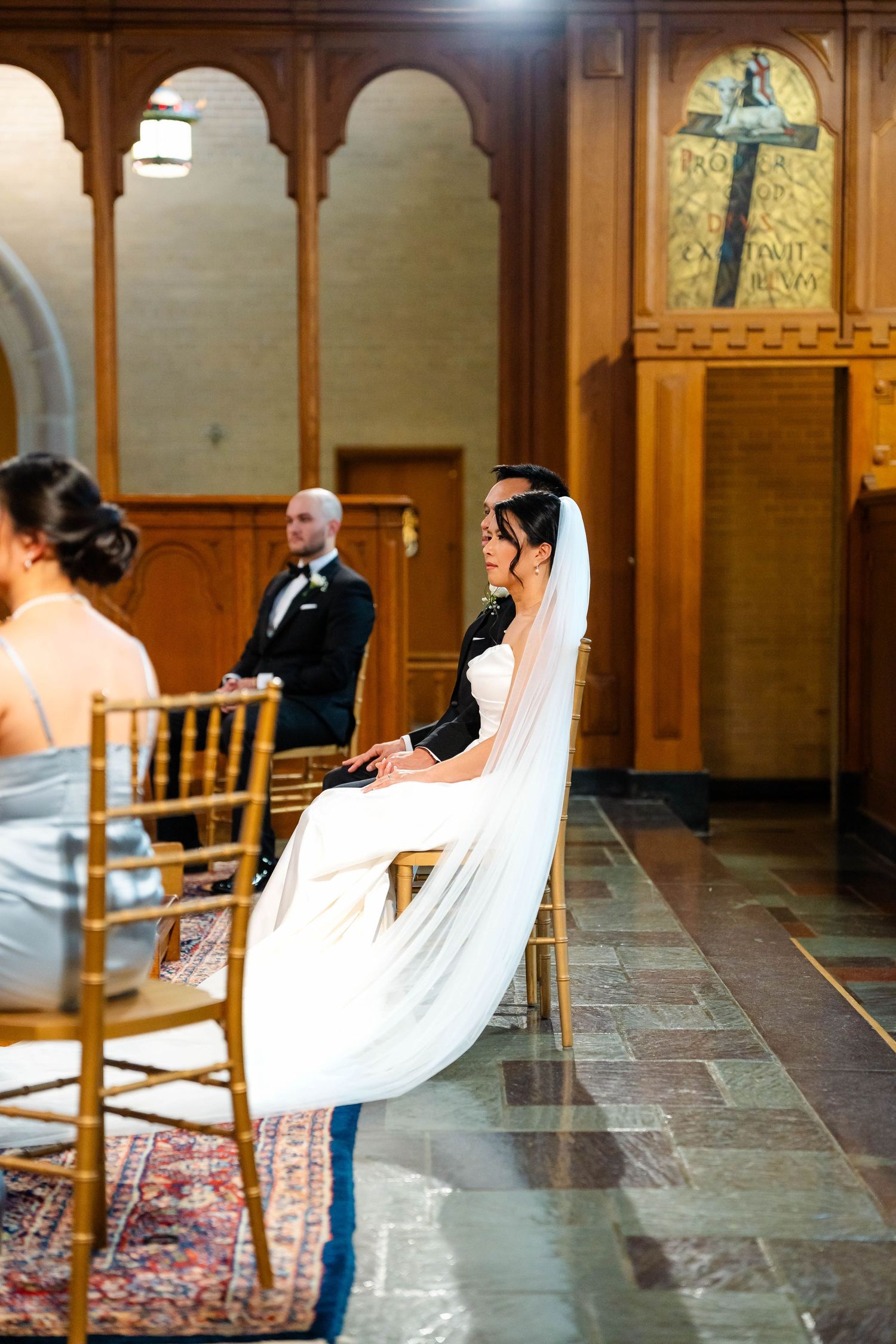 Bride in white gown and veil seated during wedding ceremony in elegant wood-paneled church interior.