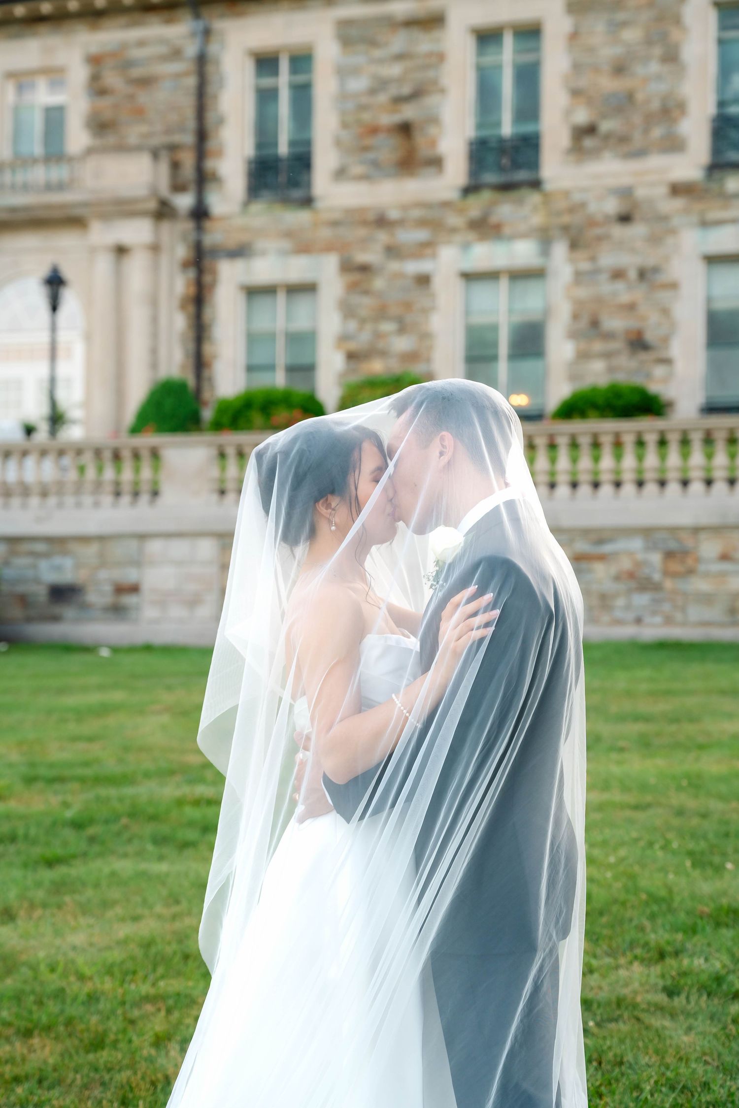 Bride and groom kiss under flowing white veil in front of stone mansion.