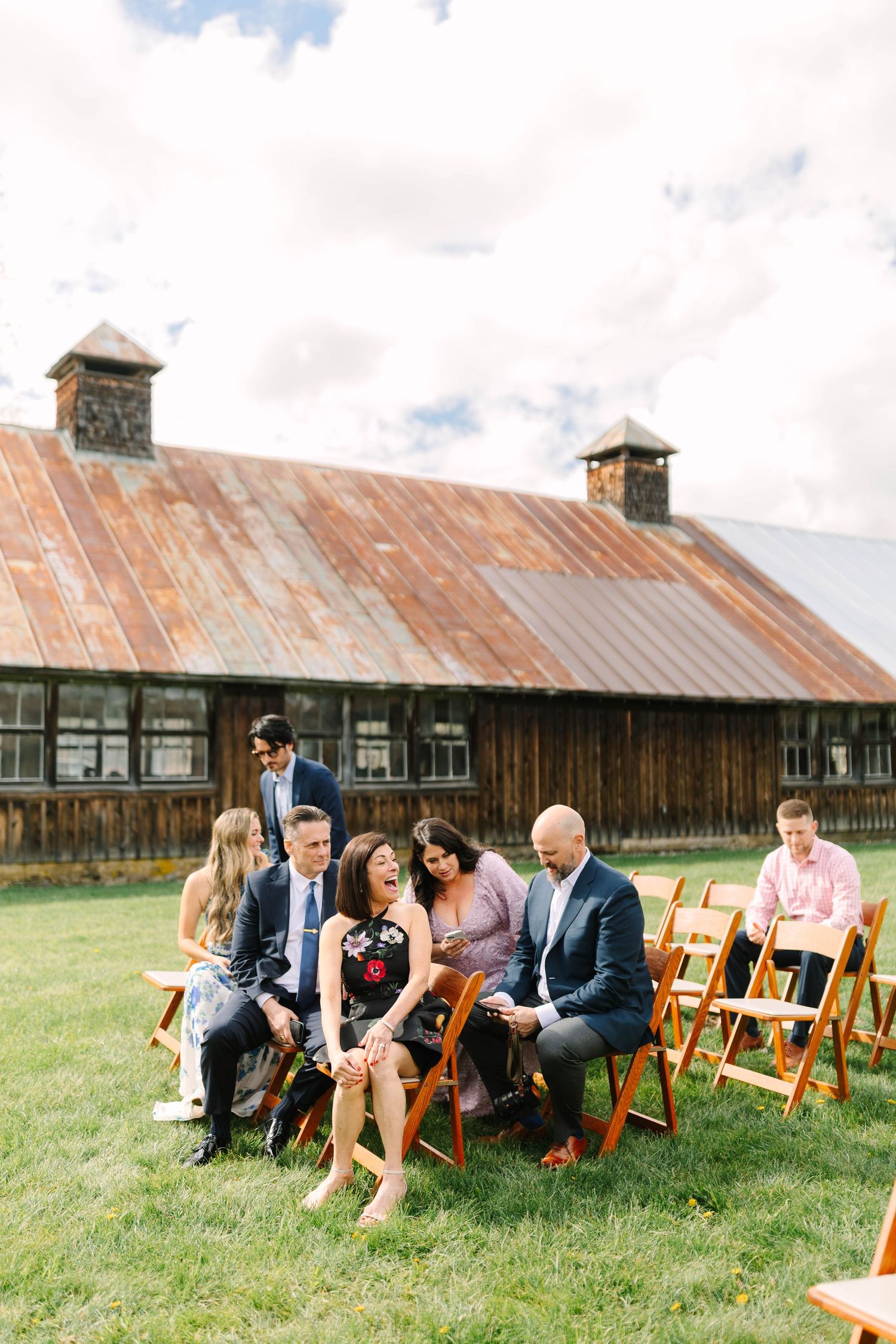 Wedding guests gather and chat on wooden folding chairs outside rustic barn venue on sunny day.