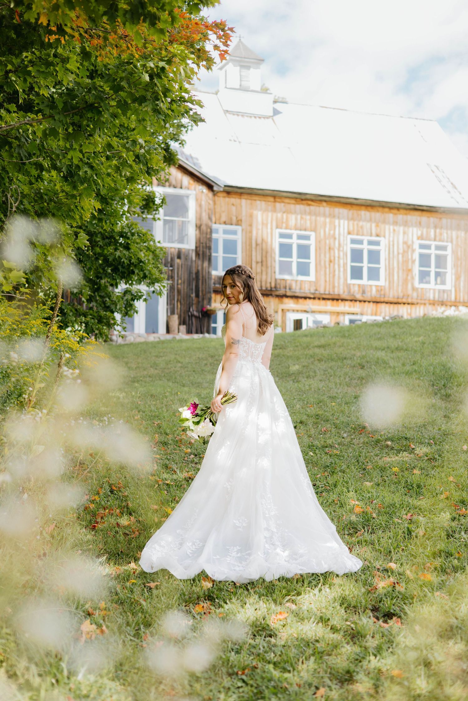 Bride in white lace gown holding bouquet stands on green grass near rustic wooden barn on sunny day.