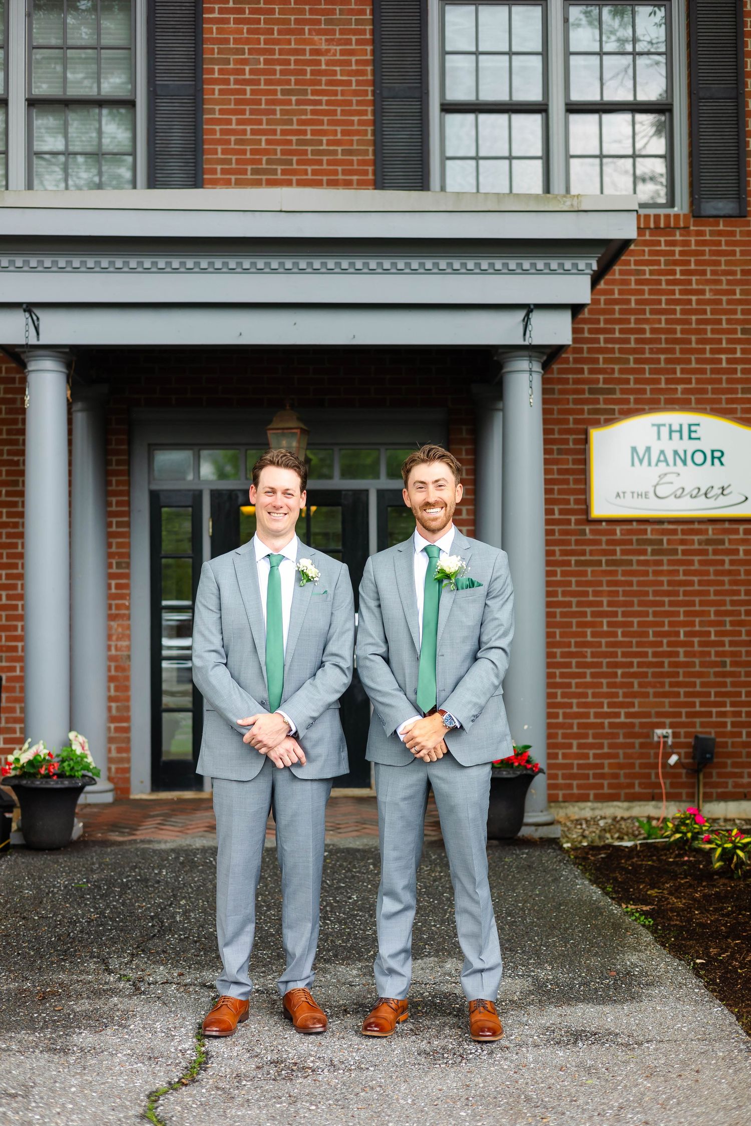 Two groomsmen in gray suits with green ties pose outside The Manor at Essex wedding venue.