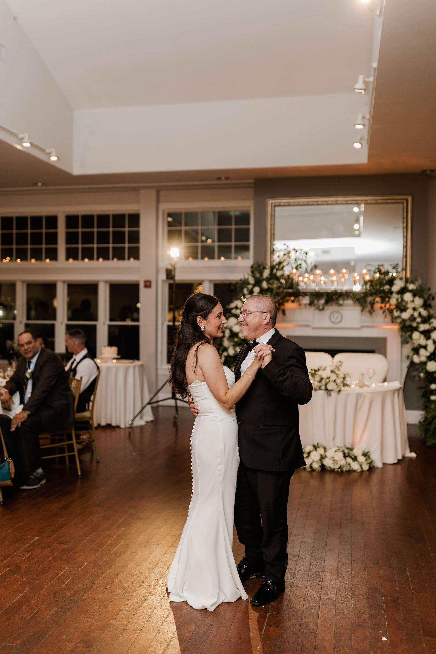 Bride and father dancing together at wedding reception with guests seated in background.