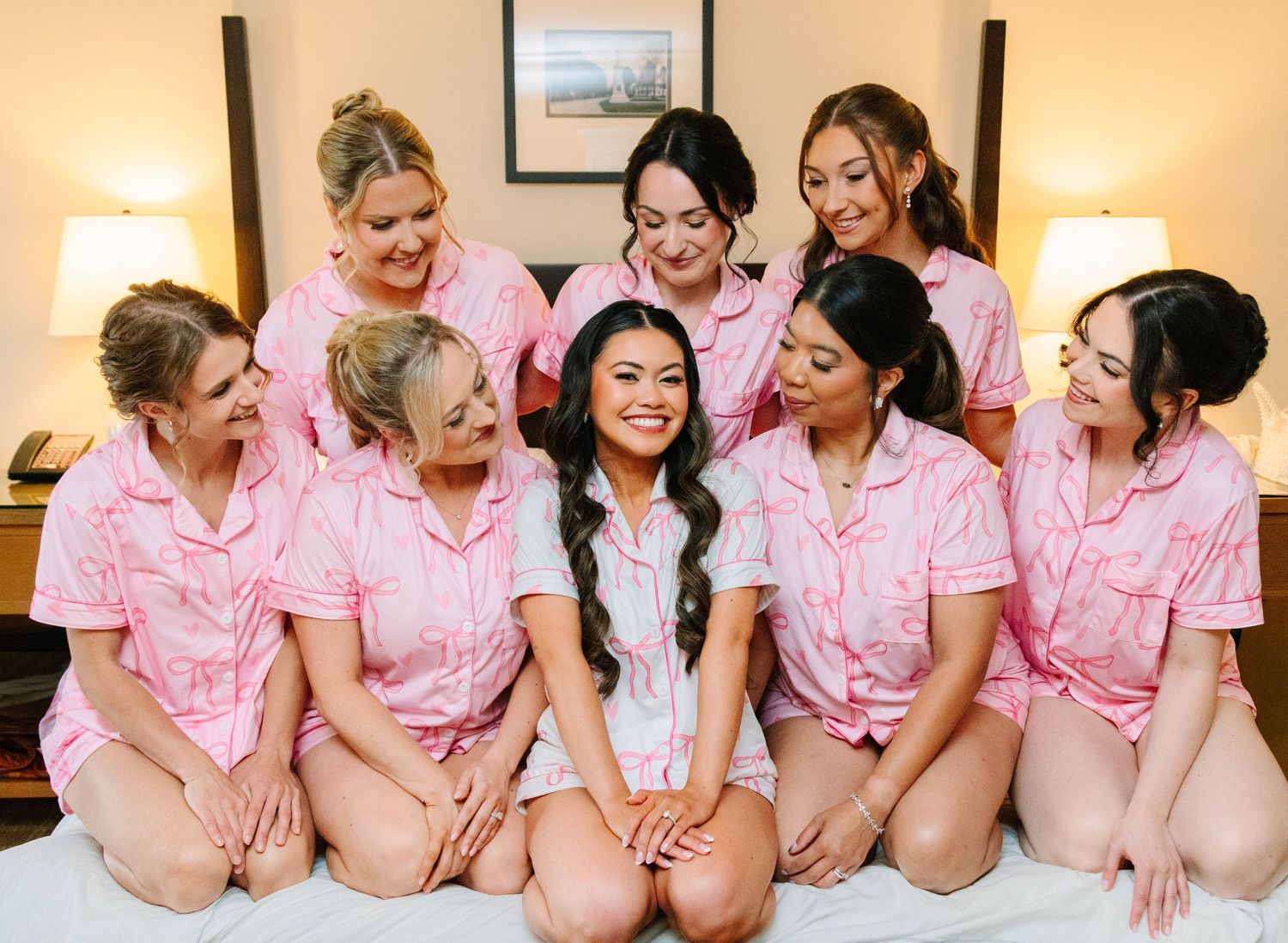 Smiling bride surrounded by seven bridesmaids in matching pink pajamas sitting on a hotel bed.