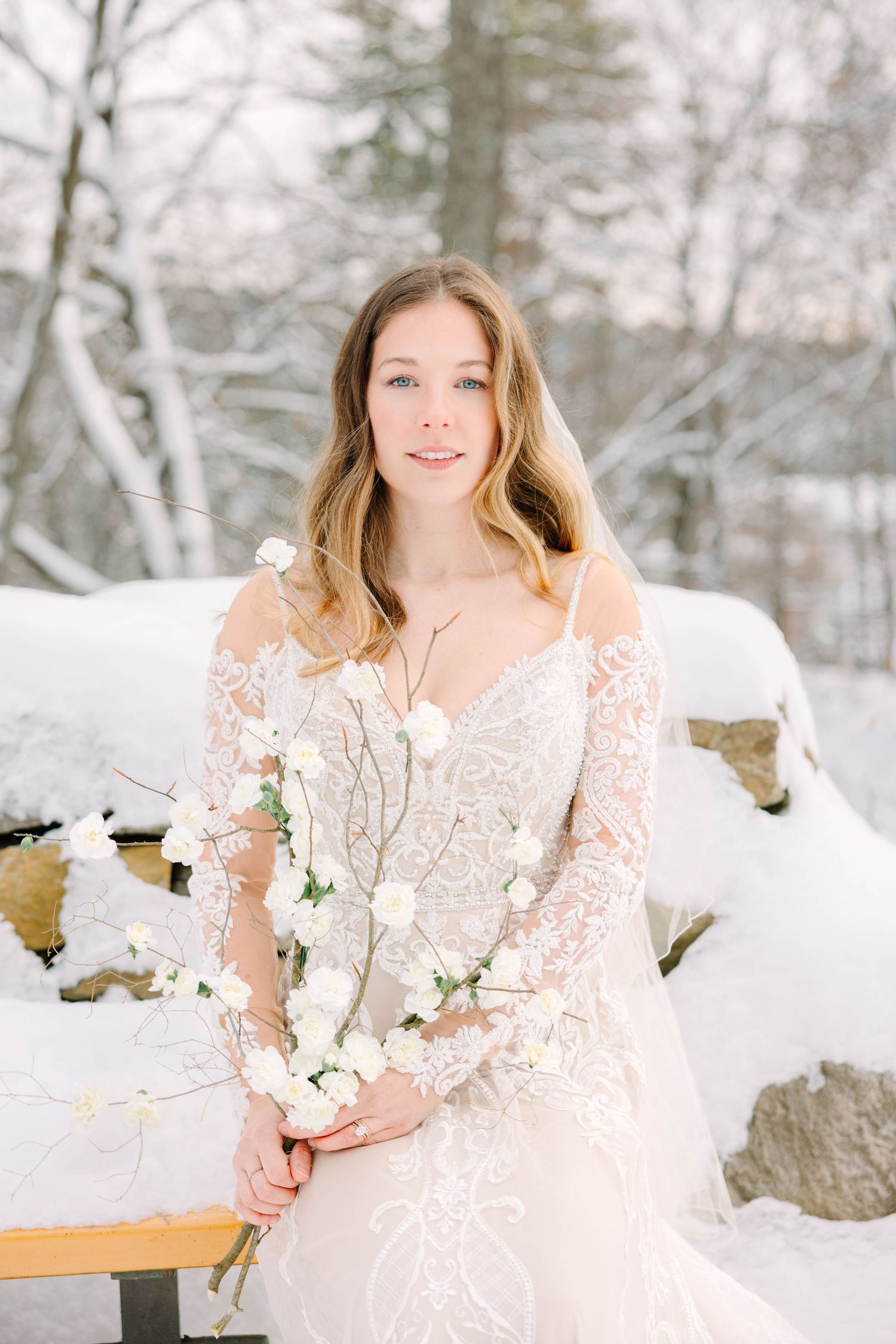 Bride in lace wedding dress holding white rose branch bouquet in snowy outdoor setting.