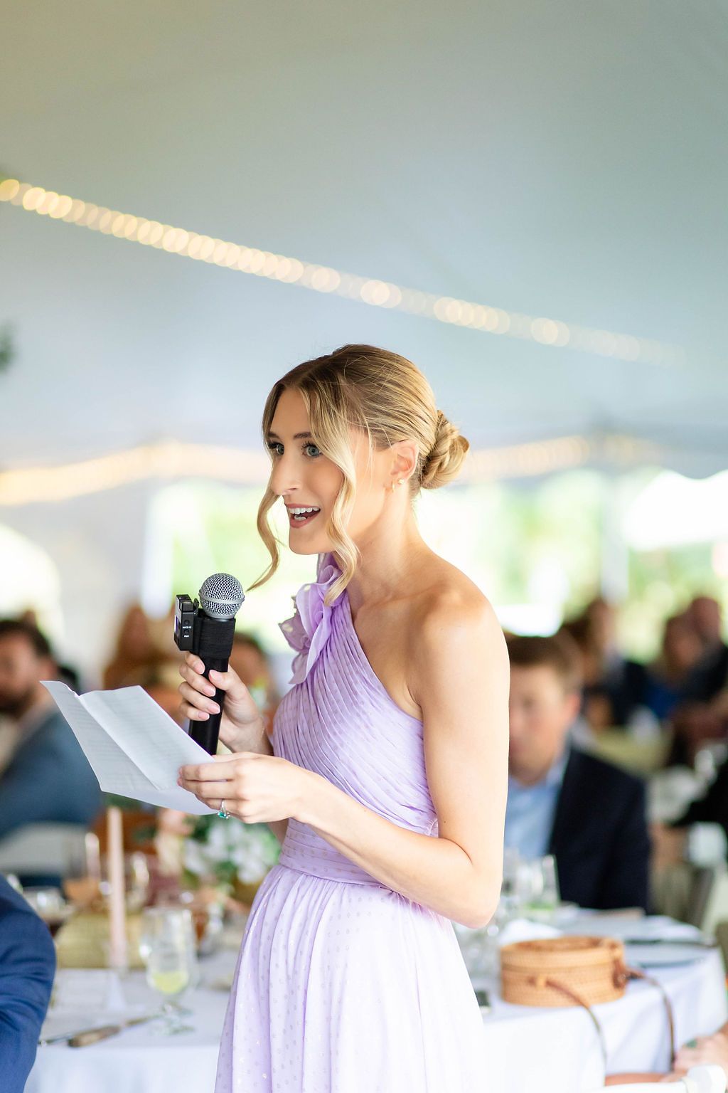 Bridesmaid in lavender dress gives a toast holding a microphone at an outdoor wedding reception.