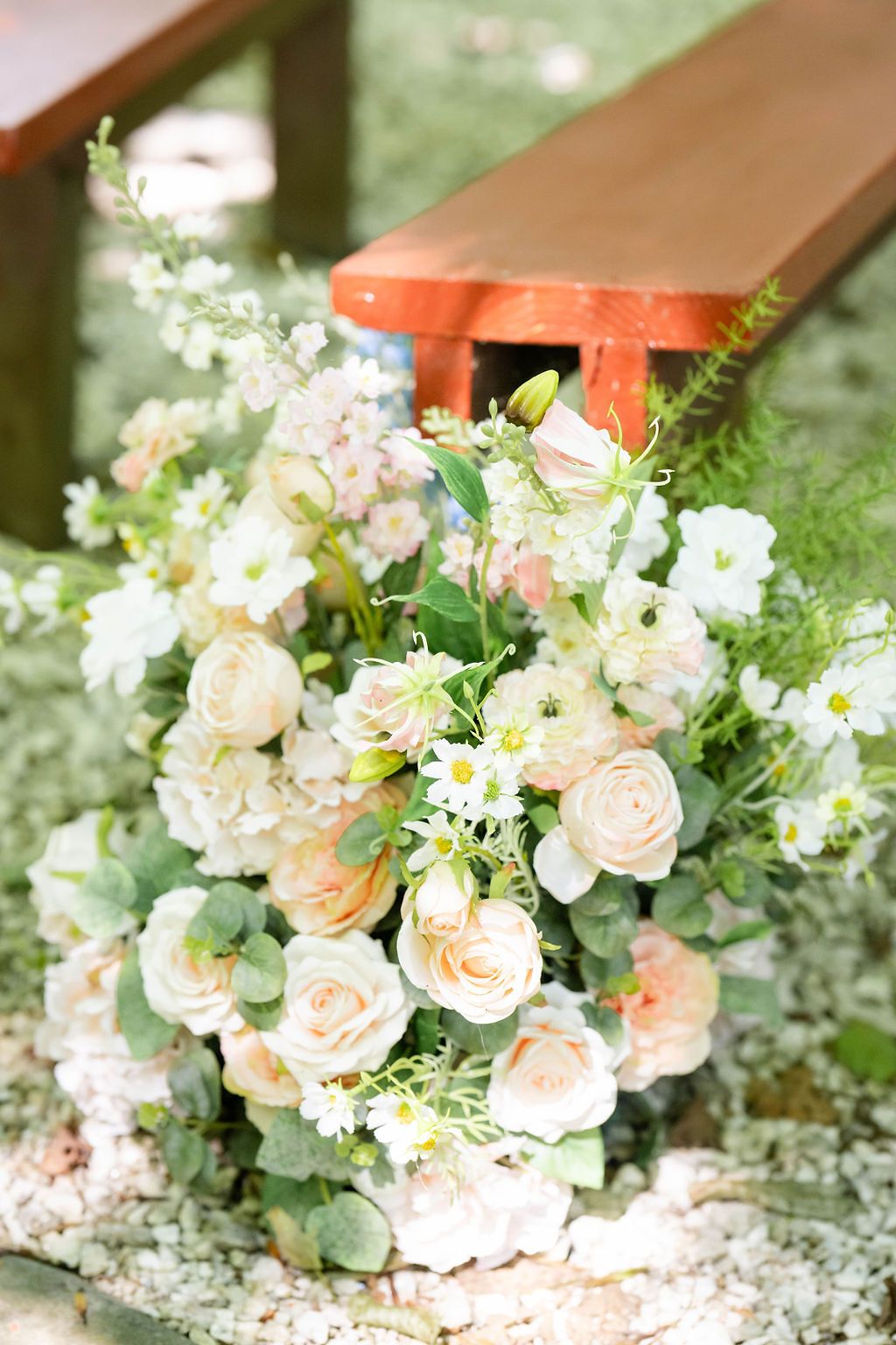 Elegant wedding floral arrangement featuring cream and blush roses with greenery beside a wooden bench.