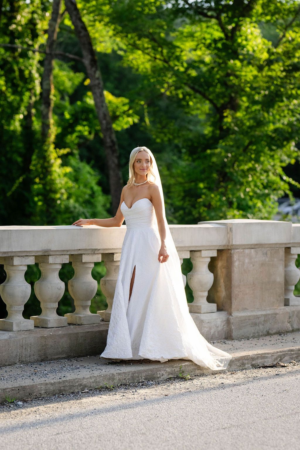 Beautiful bride in strapless white wedding gown with slit and veil posing by ornate stone balustrade outdoors.