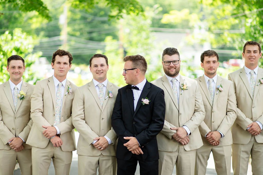 Groom in black tuxedo standing with groomsmen dressed in tan suits outdoors at a wedding.