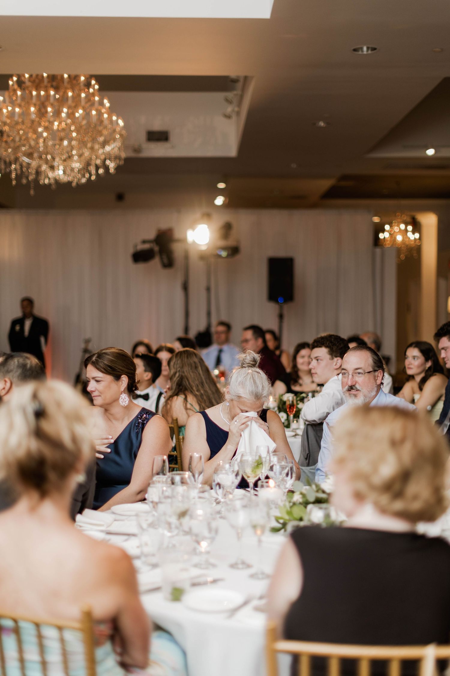 Wedding reception guests seated at elegantly decorated tables under crystal chandeliers in a grand ballroom.
