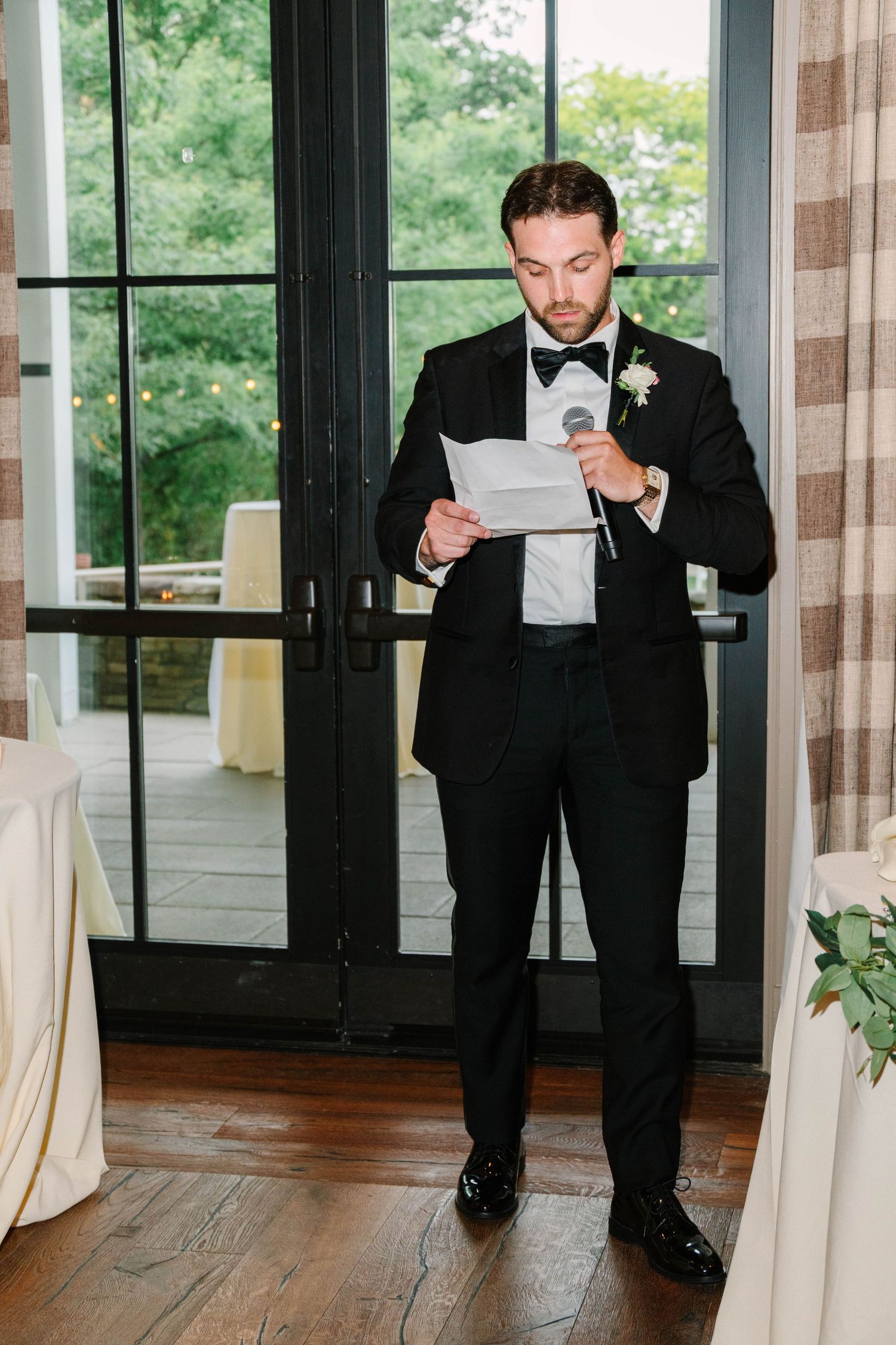 Groom in black tuxedo reads wedding speech while holding microphone at indoor reception venue.