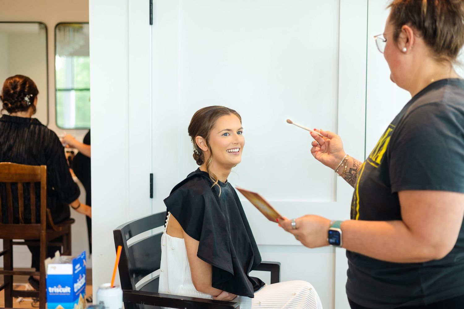 Bride smiling in salon chair while makeup artist applies cosmetics with brush during wedding prep.