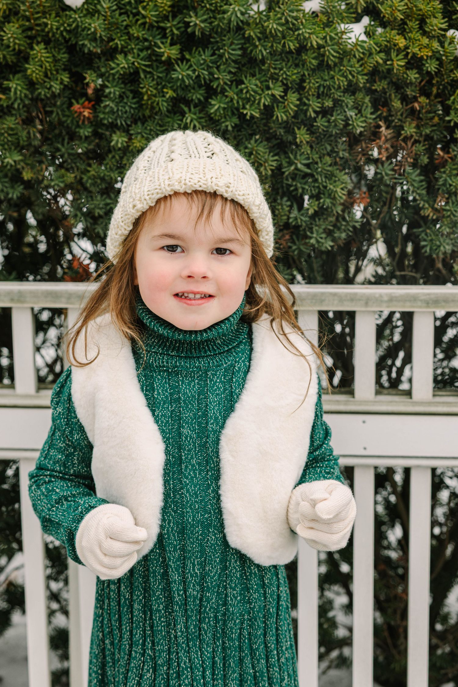 An adorable young girl in a green turtleneck dress and cream knit hat smiles outdoors in a snowy winter scene.