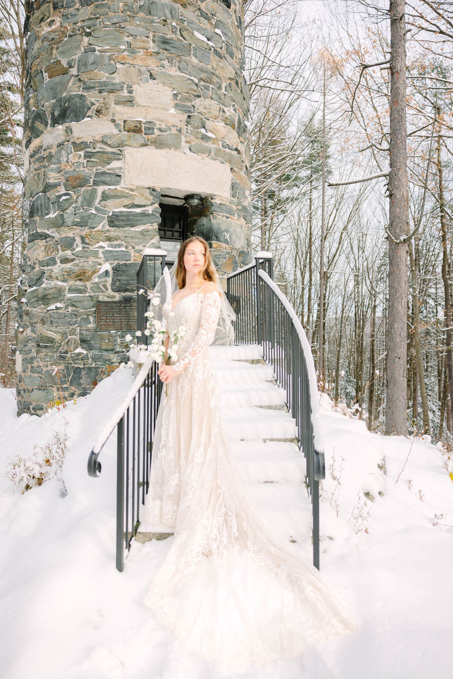 Bride in lace gown stands on snow-covered stone tower staircase in winter landscape.