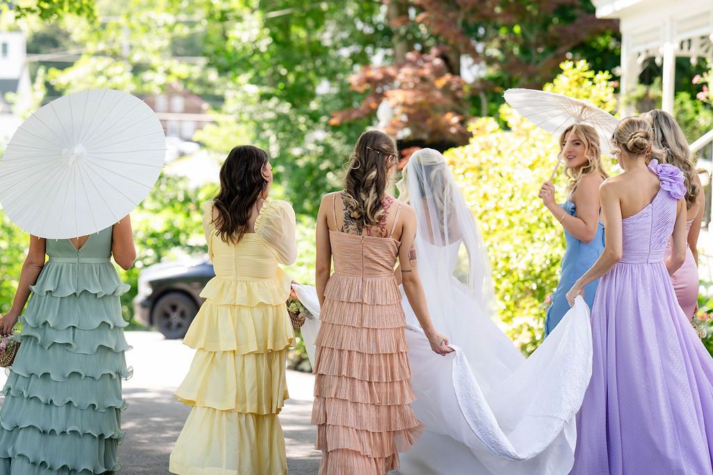 Bride in white gown and veil walks with bridesmaids in colorful pastel dresses holding parasols.