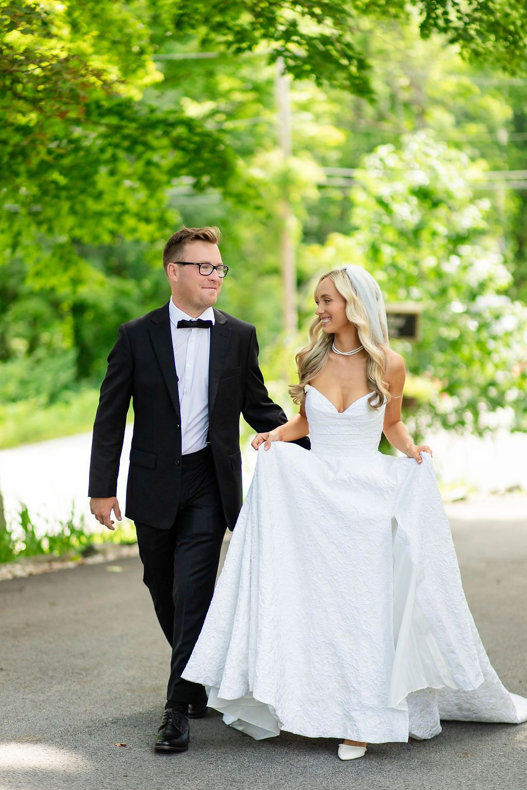 Bride and groom walking hand in hand outdoors surrounded by lush green trees on their wedding day.