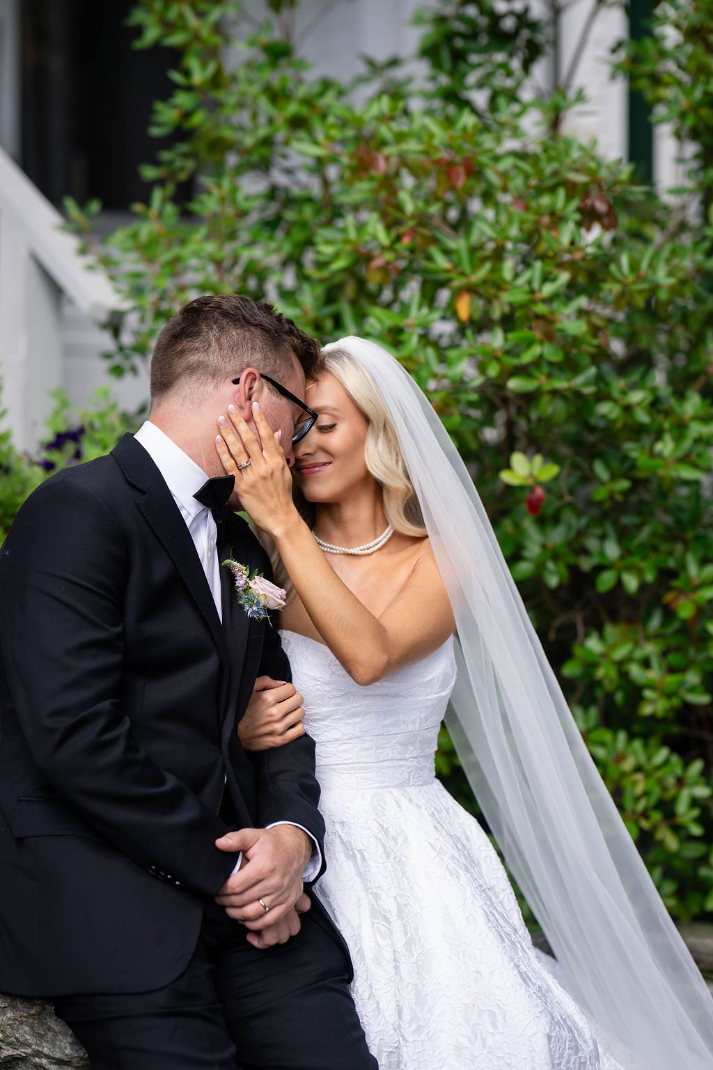 Bride in white lace gown and veil tenderly touches grooms face as they share an intimate moment outdoors.