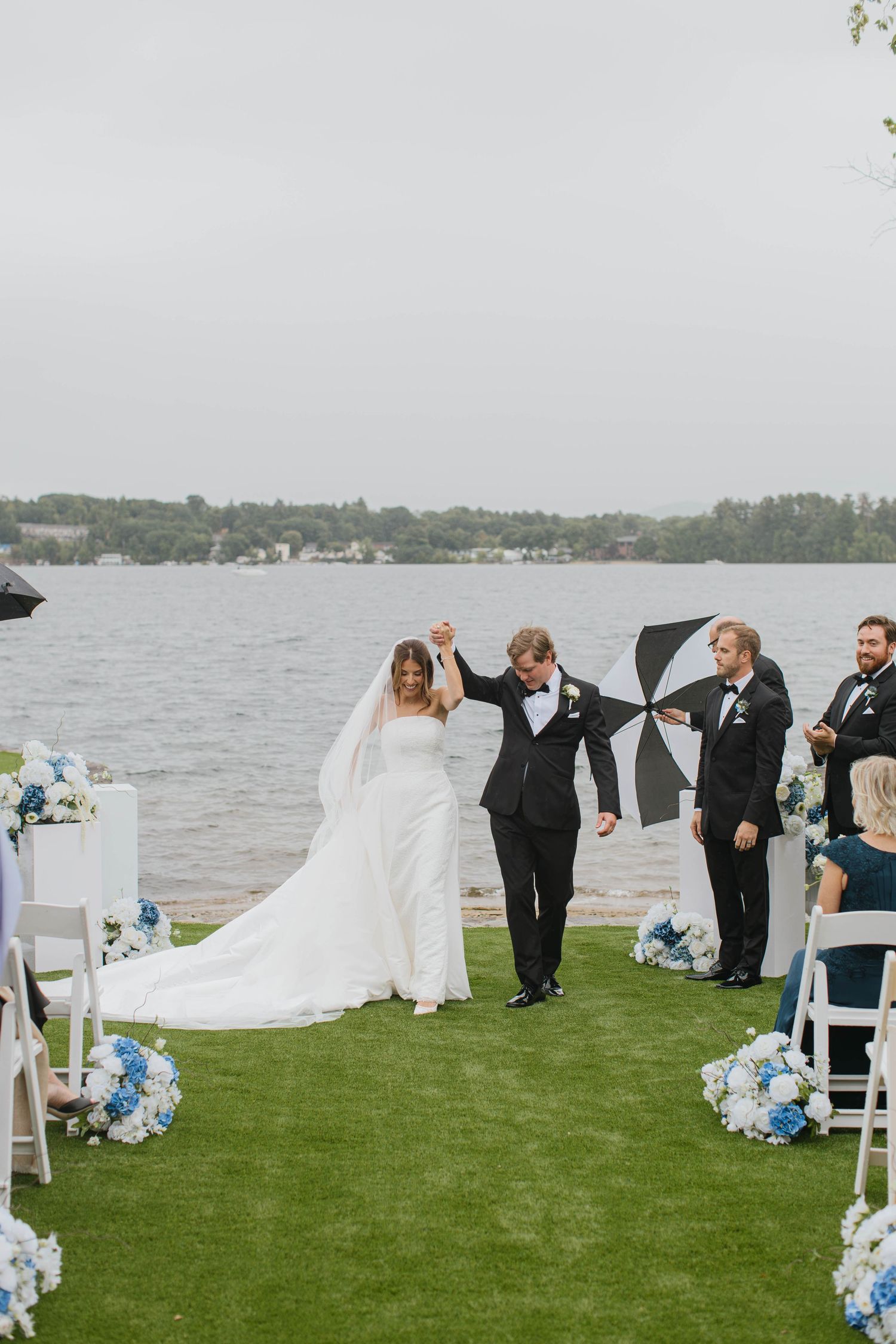 Bride and groom celebrate after outdoor lakeside wedding ceremony with blue floral arrangements on a cloudy day.