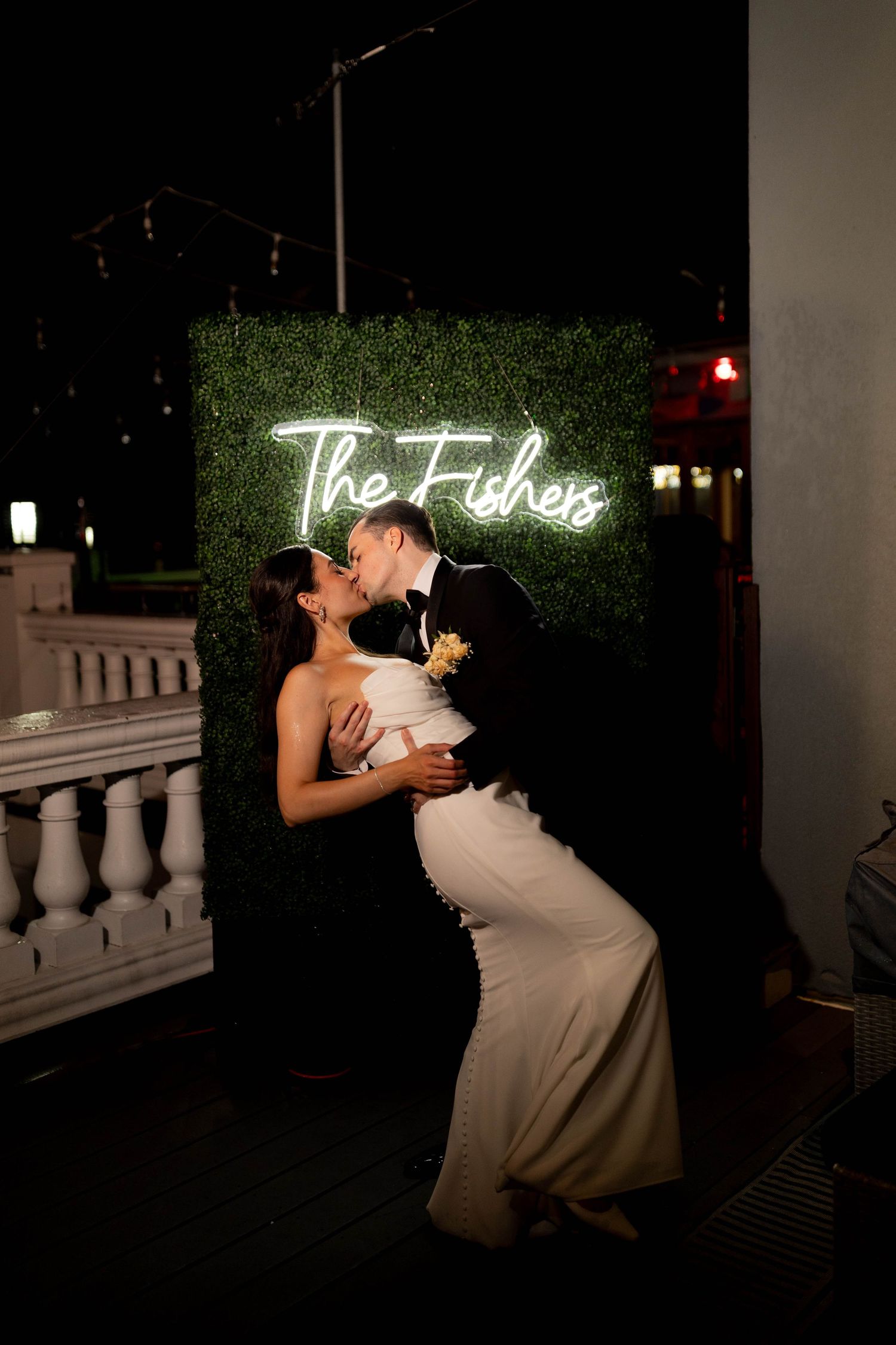 Newlyweds share a dip kiss in front of glowing The Fishers neon sign on hedge wall.