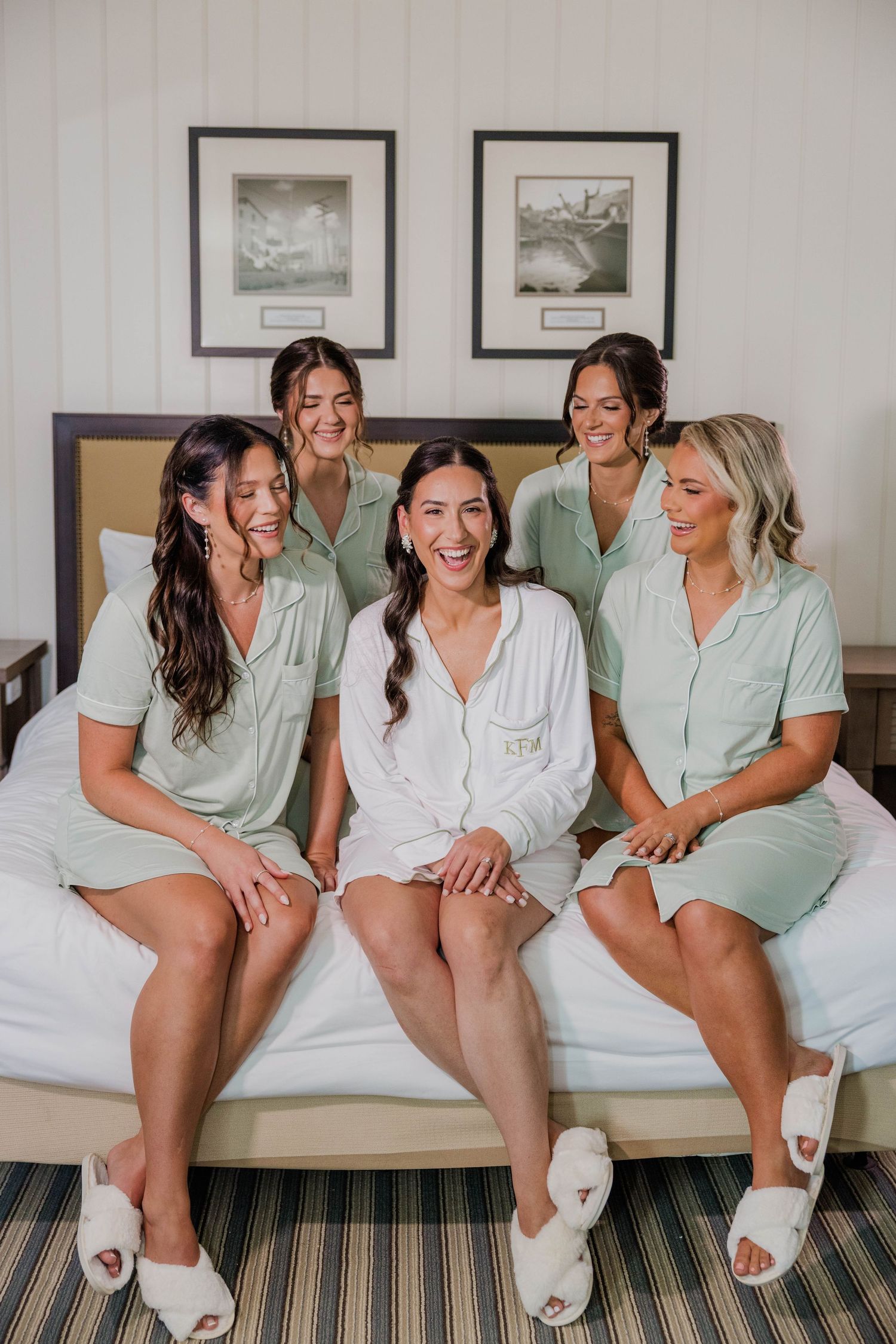 Bride in white robe sitting on bed laughing with four bridesmaids in sage green pajamas and white slippers.