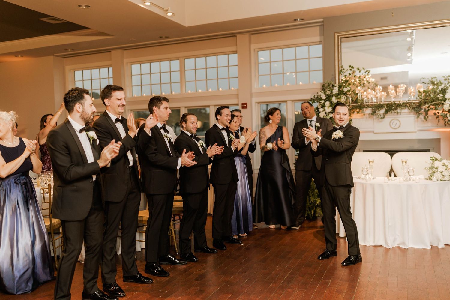 Groomsmen in black tuxedos applaud during a wedding reception in an elegant ballroom.