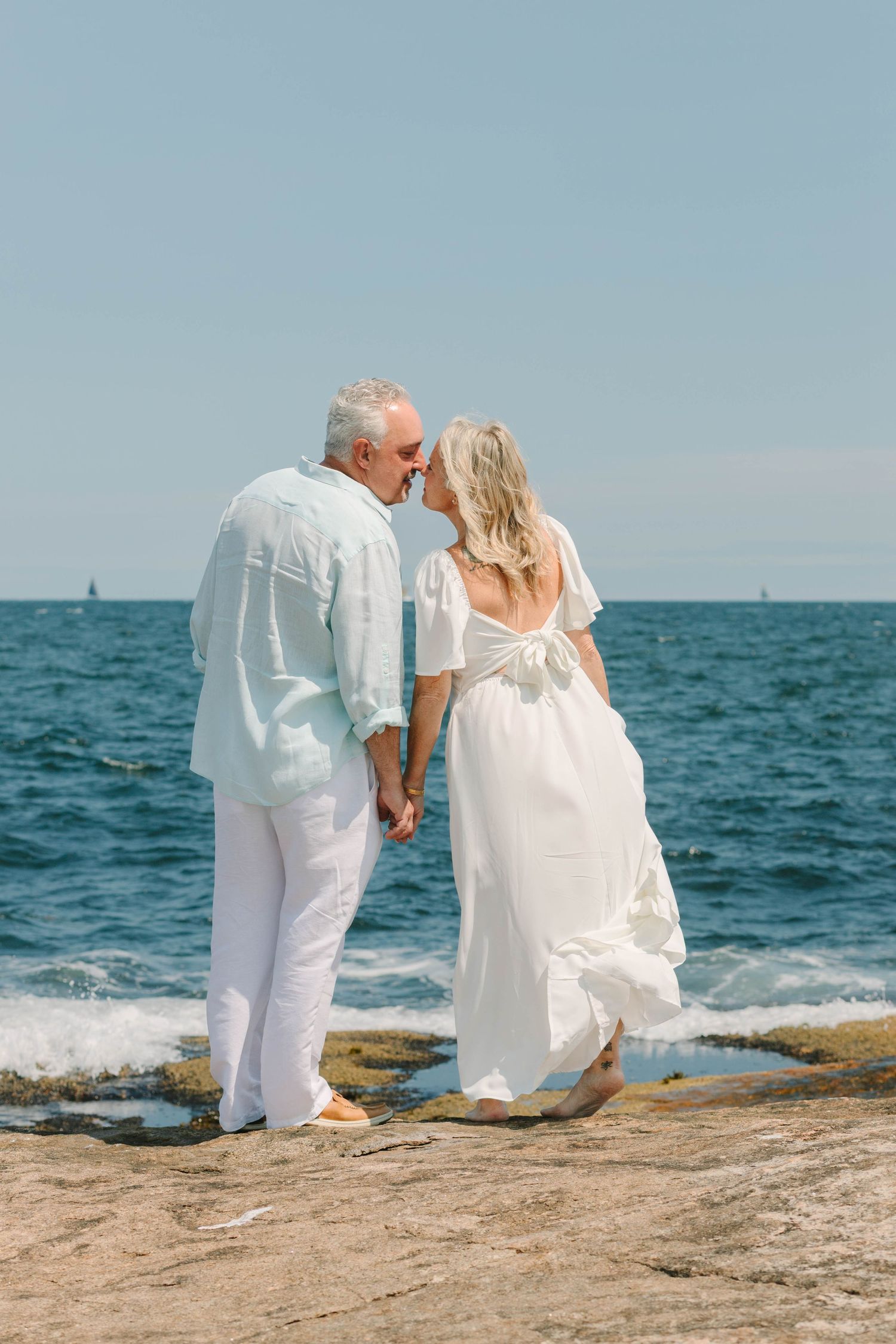 Mature couple in white attire sharing a kiss on rocky beach with ocean backdrop on a sunny day.
