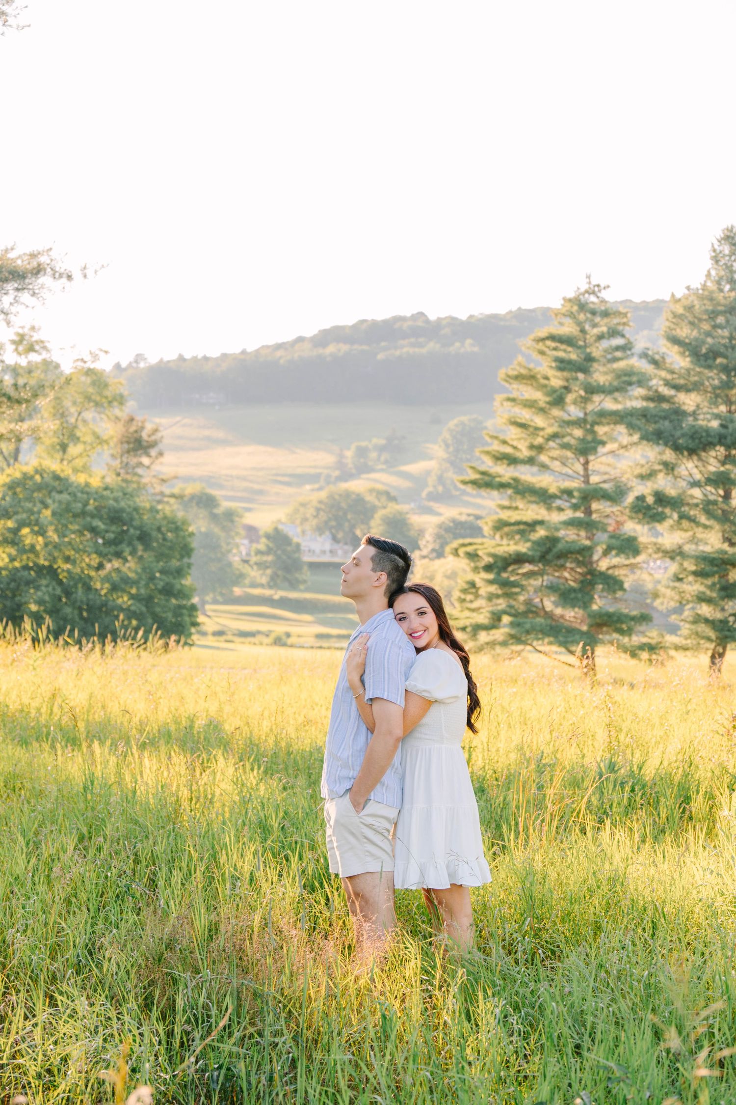 Couple embracing in a sunlit meadow with rolling hills and trees in the background during golden hour.