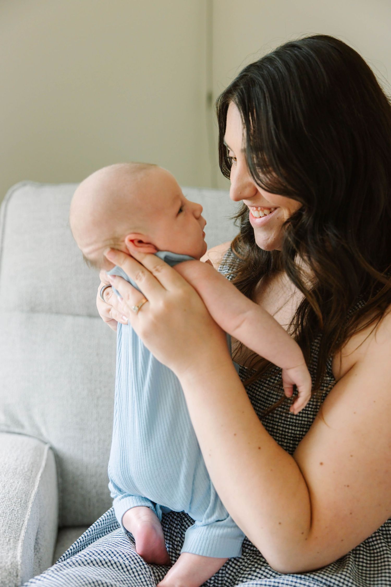Smiling dark-haired mother holding her newborn baby close while sitting on a gray couch.