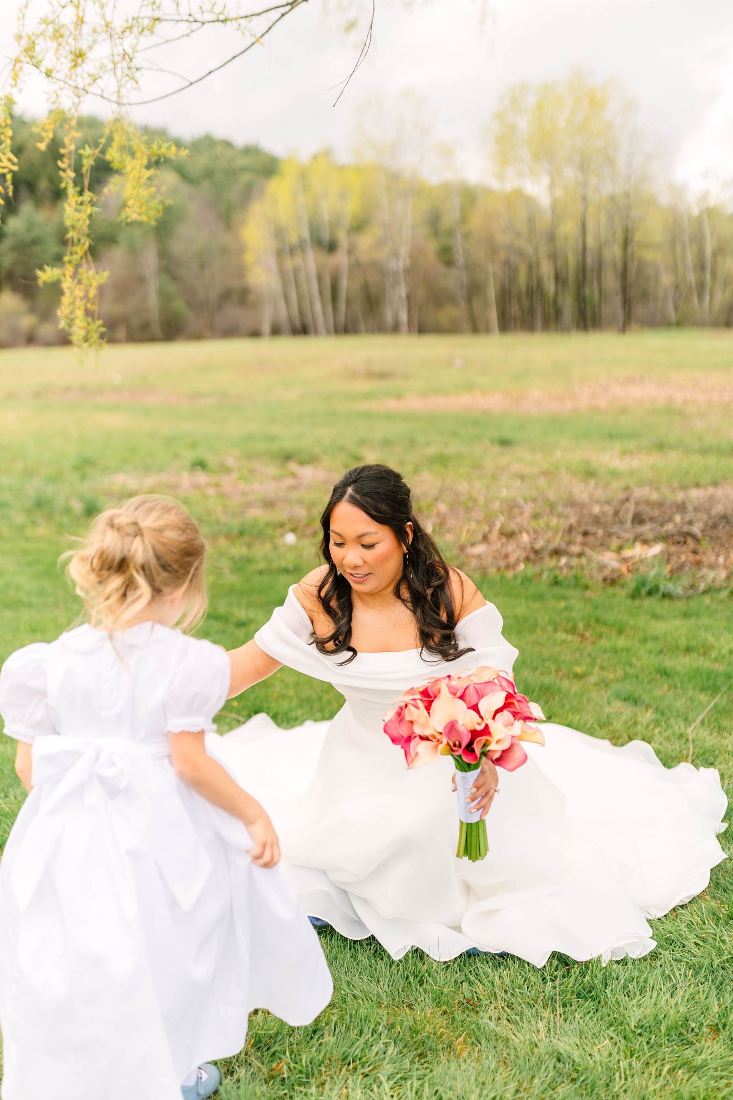 Bride in white off-shoulder gown bends toward flower girl in white dress on green lawn.